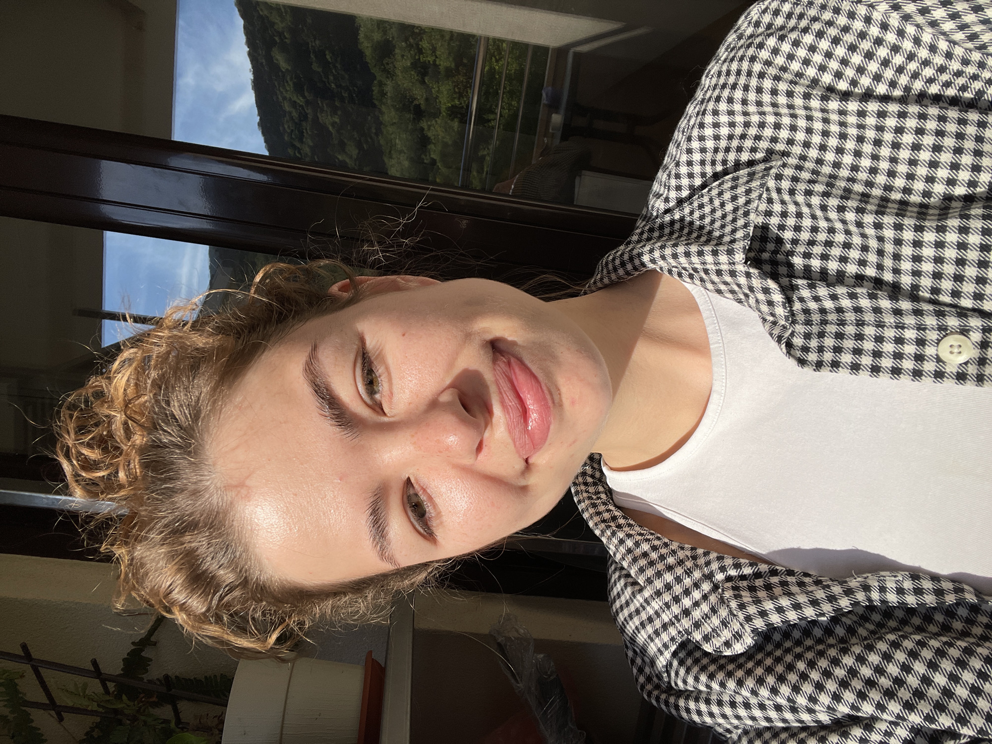 A young woman with curly blonde hair, wearing a white top and a black and white checkered jacket, is smiling and sticking her tongue out. She appears to be indoors, near a window, with a view of a green, mountainous landscape under a bright, slightly cloudy sky. The location is possibly Boreti, Montenegro, given the landscape.