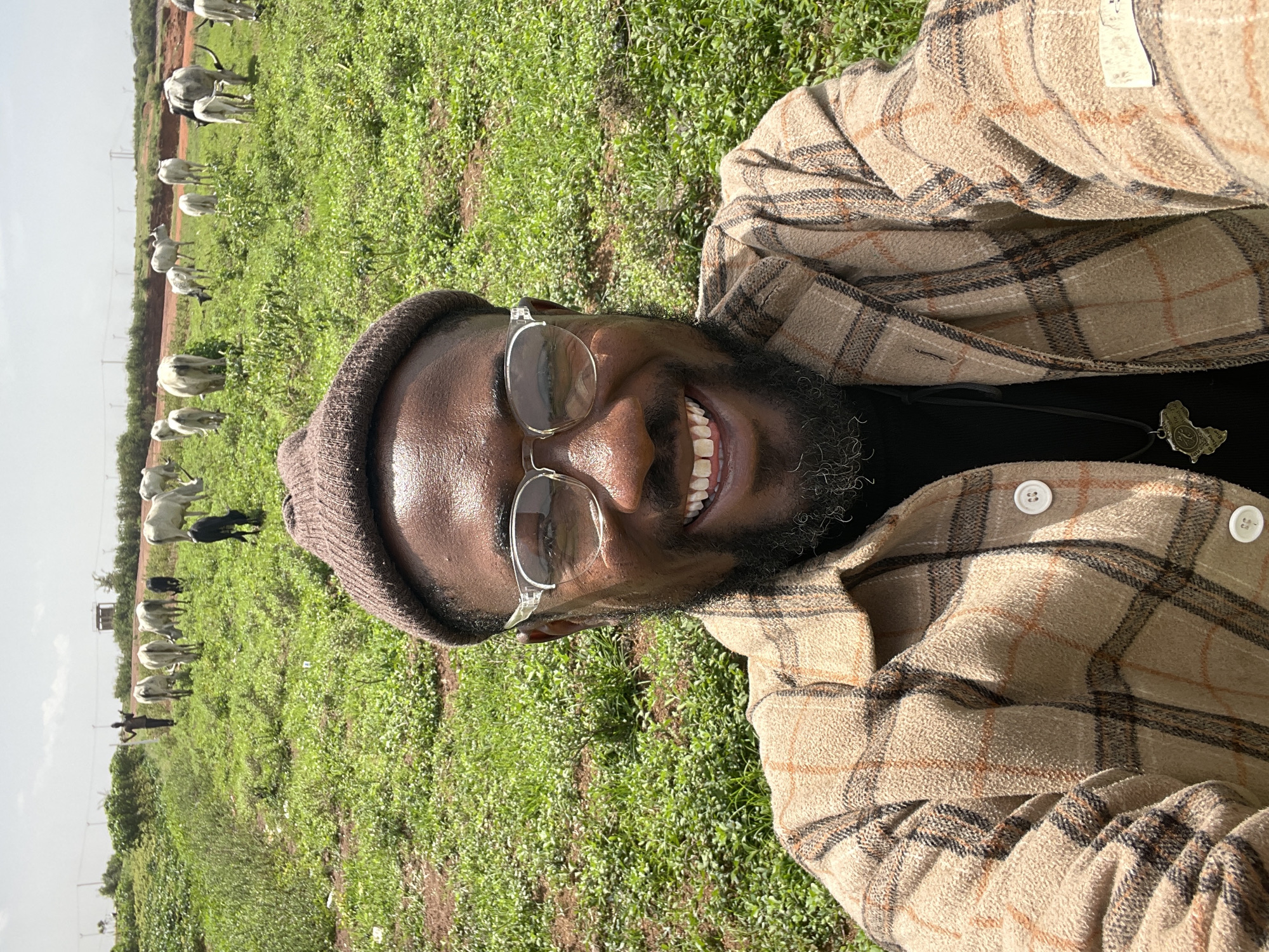 A medium shot captures a smiling Black man in a field of green grass, with cattle grazing in the background. The man is wearing a brown beanie, clear-framed glasses, a black shirt, and a brown plaid jacket with white buttons. A necklace with a map-shaped pendant is visible on his chest. The cattle are predominantly white, with a few black individuals among them. The location is speculated to be in Jos, Nigeria. The background includes a dirt path and distant utility poles. The photo appears to be a selfie.