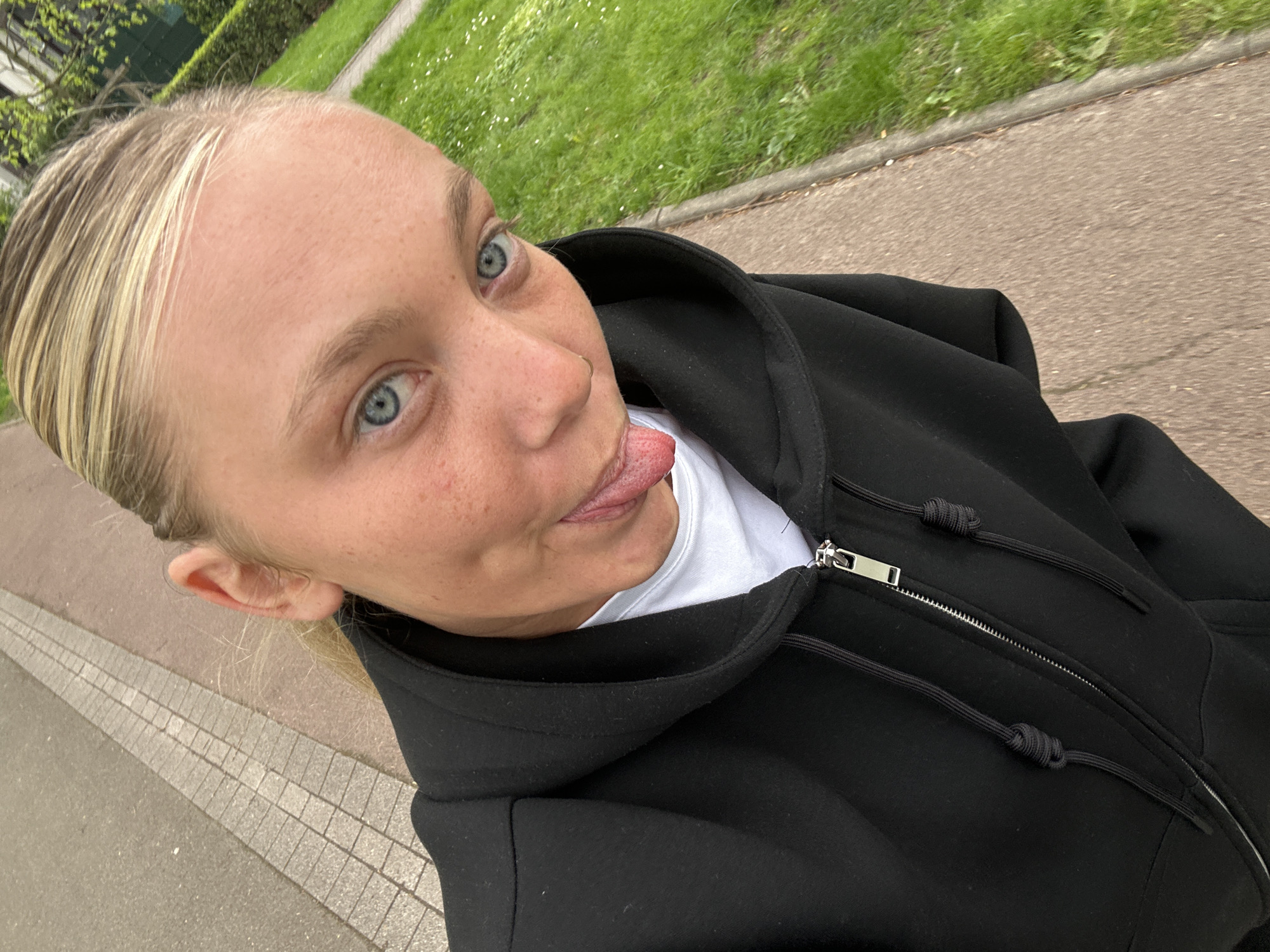 A light-skinned woman with blonde hair pulled back is sticking her tongue out and looking towards the camera. She has freckles and blue eyes. She is wearing a black hooded sweatshirt over a white shirt. The background consists of a paved walkway, a curb, and a grassy area. Based on available information and visual cues, the location is consistent with Noisiel, France.