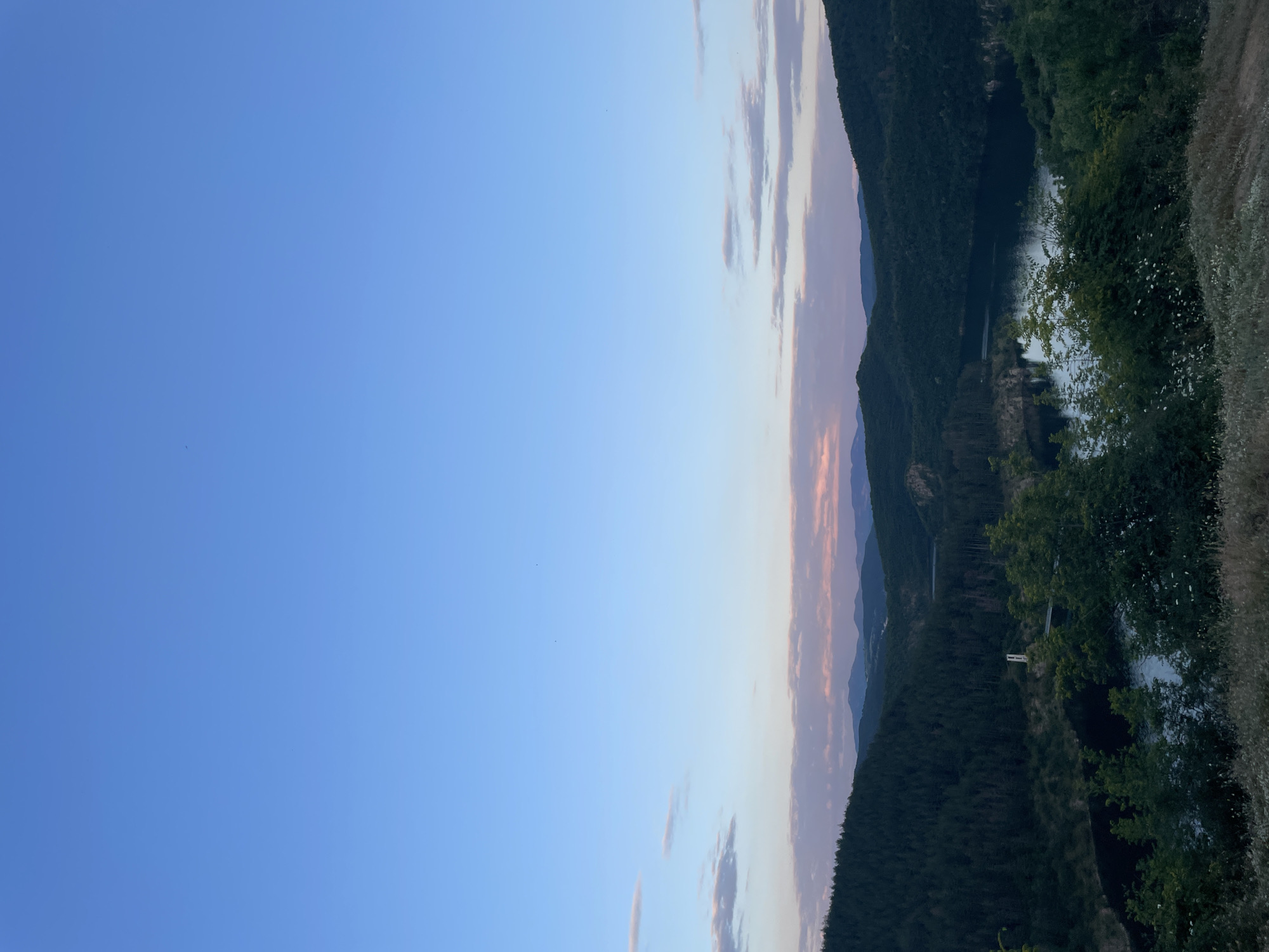 The image depicts a panoramic view of a serene natural landscape at dusk, likely in or near the city of Sukhovo, Bulgaria. The sky is a gradient of pale blue to a soft pink and lavender near the horizon, with a few wispy clouds. Rolling hills covered in dense, dark green coniferous forests dominate the midground.
In the center of the image, a dark, still body of water, possibly a lake or reservoir, is nestled within the valleys. A thin, white vertical structure, likely a part of a bridge or a tower, is visible on a slope overlooking the water. The foreground is a gentle slope covered in dry grasses and scattered small white wildflowers, suggesting an elevated vantage point. The overall atmosphere is peaceful and tranquil, capturing the quiet beauty of nature as daylight fades.