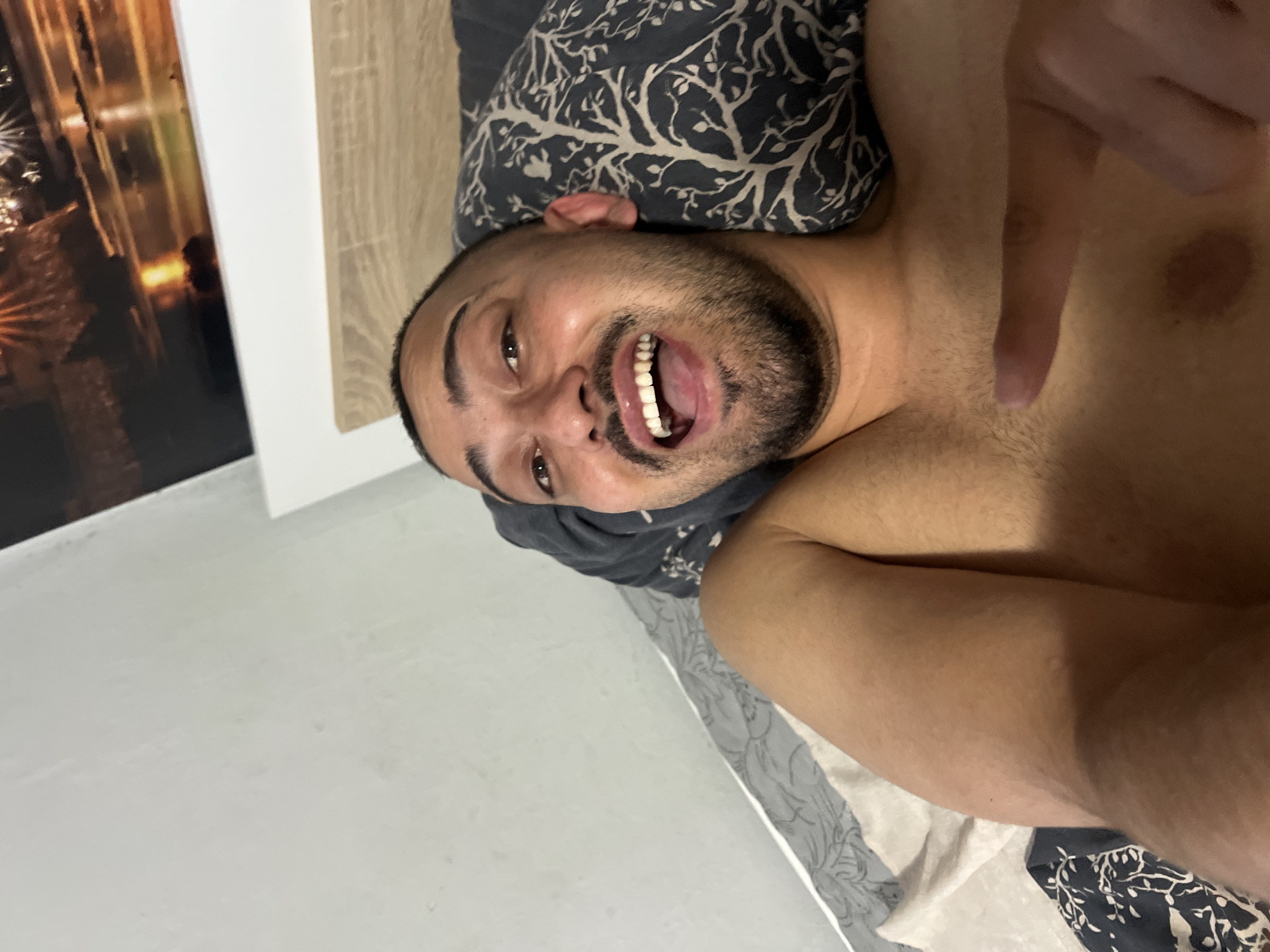The image shows a close-up, tilted selfie of a man lying in bed. The man has dark hair and a beard and is wearing a dark t-shirt. He is smiling broadly with his mouth open, revealing his teeth. His eyes are looking directly at the camera. The background includes a pillow with a dark, abstract pattern and a wooden headboard. To the left, a white wall is visible, with a framed picture of what appears to be city lights at night. The lighting suggests it might be nighttime or an indoor setting with artificial light. The man's expression conveys amusement or excitement. No visible text is present in the image.