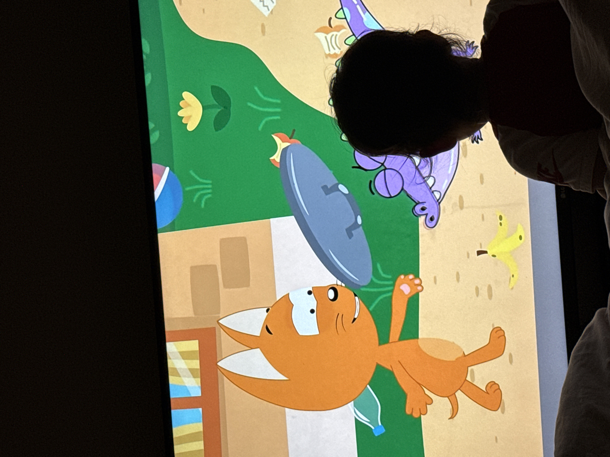 The image shows a child watching a cartoon on a television screen. The cartoon depicts an orange cat character interacting with a purple alligator character. The scene is set outdoors with elements such as grass, a flower, and a banana peel. The child is silhouetted, with their back to the camera, and is wearing a t-shirt. The television is dark around the edges, indicating it's the main light source. The setting appears to be a living room or bedroom. There is no information in the image to suggest the location is Gwangju, South Korea.