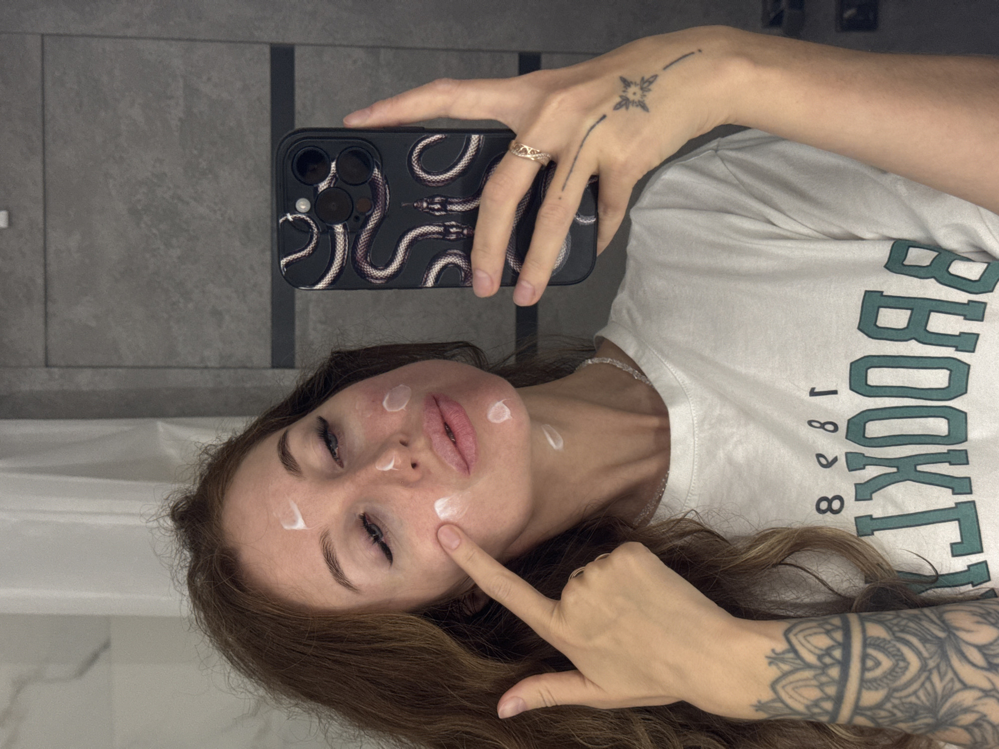 The image shows a woman taking a selfie in what appears to be a bathroom. She has her hair down and is wearing a white t-shirt with green lettering. There are several dots of cream on her face, and she is pointing to her cheek with her left index finger. She is holding a smartphone with a snake design on the back in her right hand. Her left arm has a tattoo with a mandala design, and her right hand has a ring on her finger and a small tattoo on the back of her hand. The background appears to be a tiled wall. The image does not provide any information that would allow for its location to be identified as Chaikovsky, Russia.