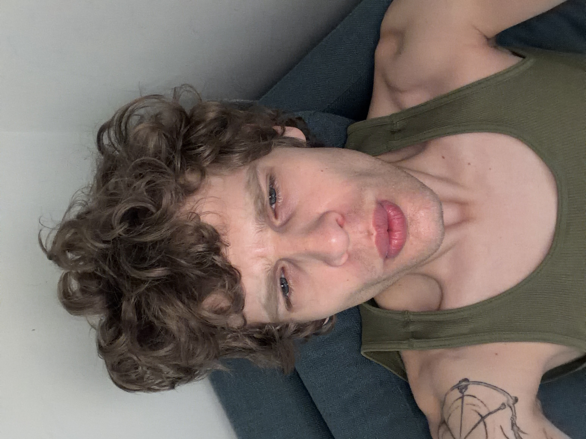 A man with curly brown hair and blue eyes is lying on a blue surface. He is wearing a green tank top, and a tattoo is visible on his shoulder. The background is a plain white wall. The image is taken from an overhead perspective. There is no indication of Milan, Italy.