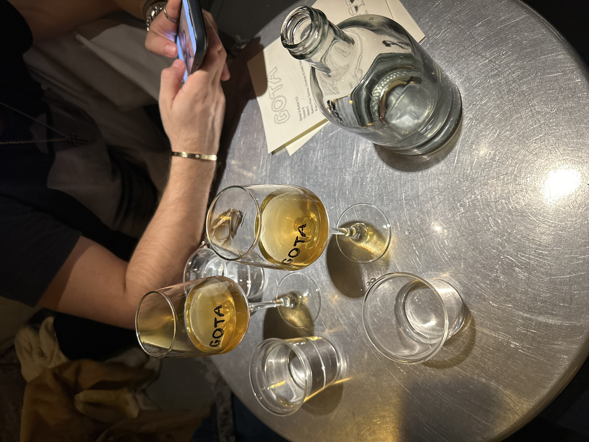 The image captures a high-angle view of a round, brushed silver table at "GOTA," a restaurant in Madrid, Spain, as indicated by the menu. A person is holding a smartphone, possibly taking a picture of the scene. The table is set with an empty bottle, two wine glasses filled with a light-colored liquid, two empty glasses, and a menu. The lighting is somewhat muted, emphasizing the metallic sheen of the table. The person is wearing a dark shirt, a silver bracelet, and a necklace, and is seated at the table.