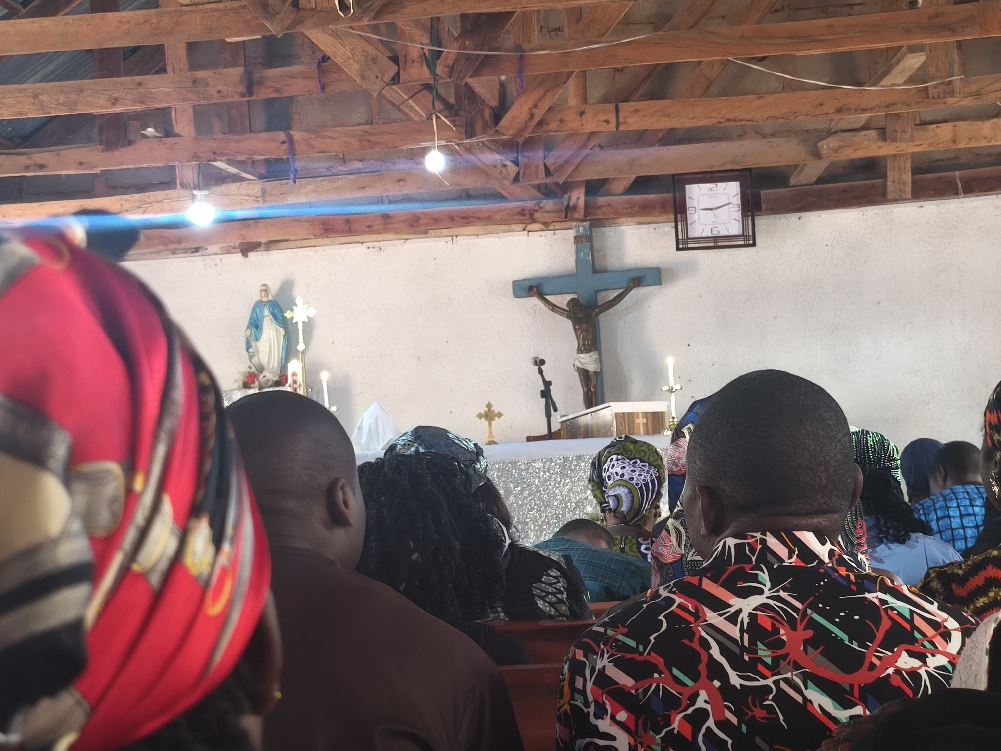 The image depicts the interior of a church in Nabatum, Nigeria. The church has a simple altar with a blue crucifix and a statue of Mary. A wall clock hangs to the right of the crucifix. The roof structure is made of exposed wooden beams. The congregation consists of people dressed in various colorful African prints and head coverings. They are seated in rows of pews. The focus is on the backs of the congregants facing the altar. The lighting is a combination of natural light and artificial light from ceiling fixtures.