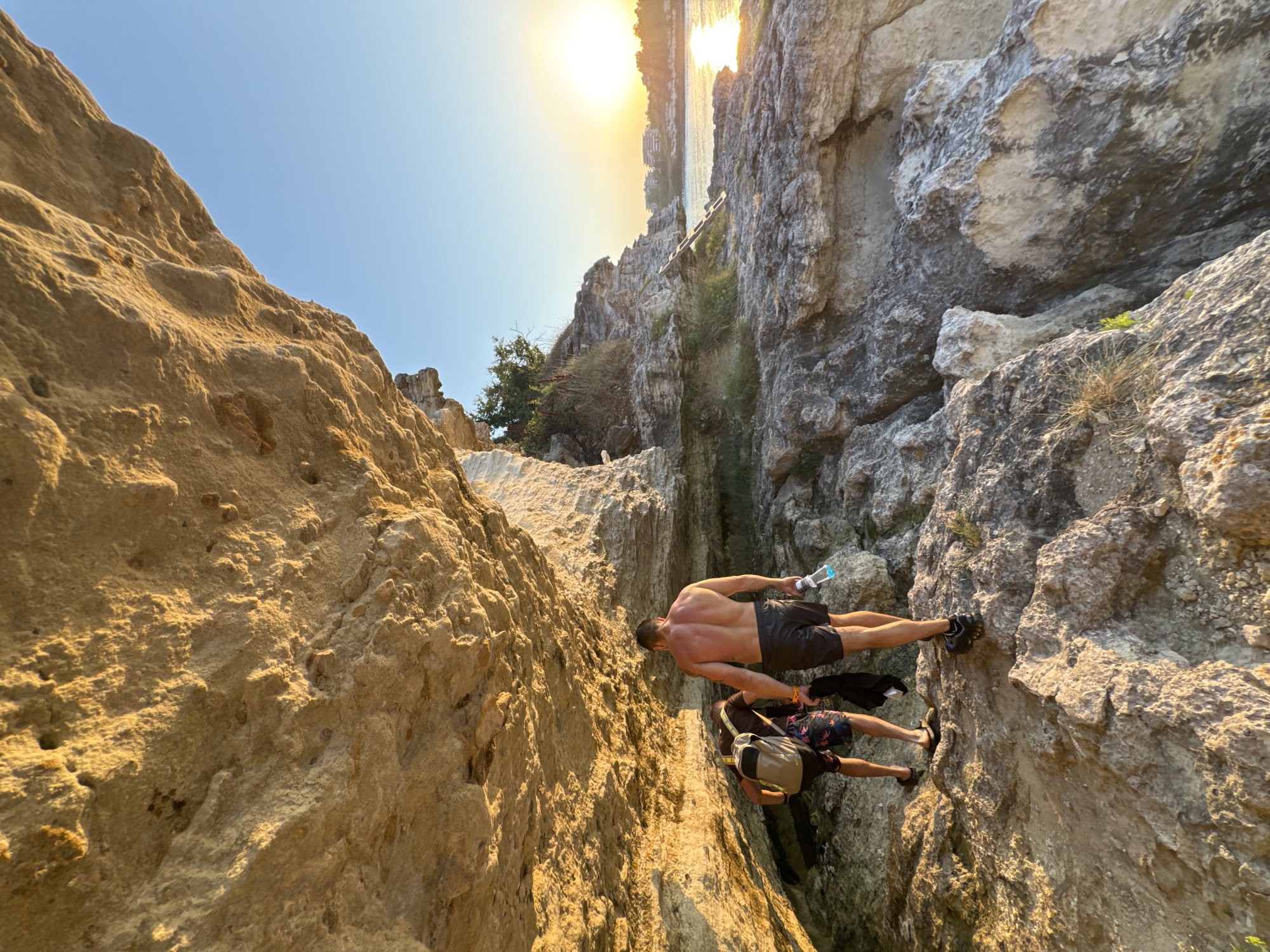 Two men walk under a rocky overhang near the coast in Tsarevo, Bulgaria. The man in the foreground is shirtless and wears black shorts and athletic shoes. He holds a water bottle. The man behind him wears patterned shorts, a dark t-shirt, and a backpack. He also carries a black garment. The terrain is rocky and uneven, with patches of dirt and sparse vegetation. In the background, the sun sets over the Black Sea, casting a warm glow on the water and distant buildings. The rocky formations suggest a natural coastal landscape.