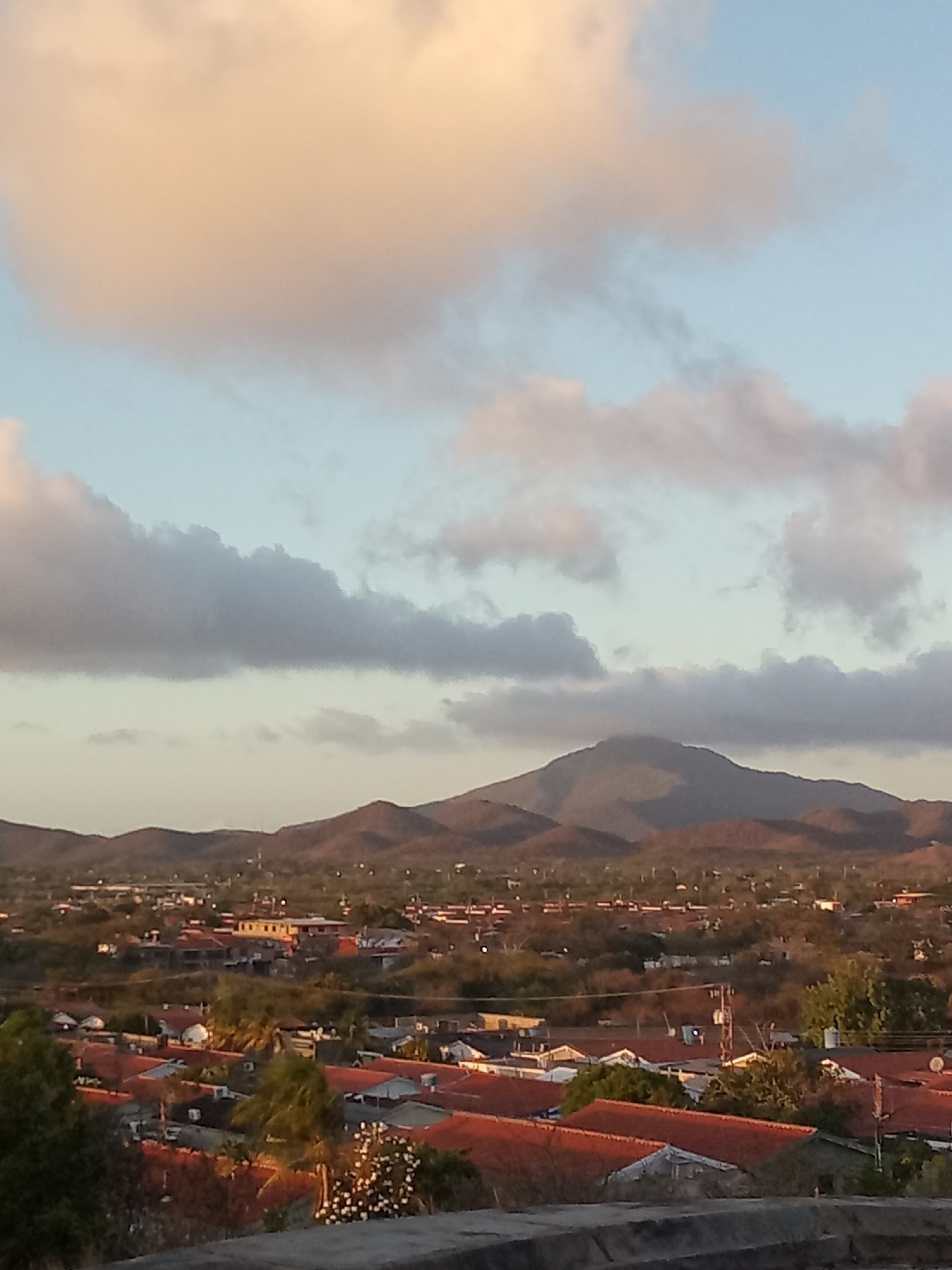 The image presents a view of the city of Juan Griego, Venezuela, under a partly cloudy sky. In the foreground, there is an elevated platform or ledge. Below the ledge, the cityscape unfolds with numerous buildings featuring predominantly red tile roofs intermixed with green vegetation. In the mid-ground, beyond the buildings, lies a series of rolling hills, with a prominent mountain dominating the horizon. The sky is pale blue, dotted with white and light gray clouds, some tinged with a warm, golden hue from the setting or rising sun. No people or specific actions are visible in the image.