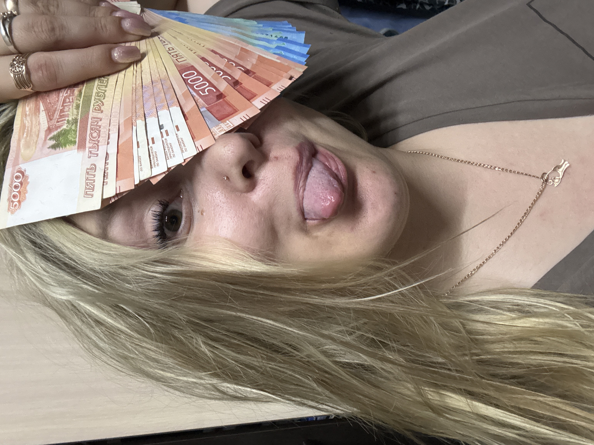 A fair-skinned person lies on their side, holding a fan of Russian ruble banknotes in front of their face, obscuring part of their forehead and right eye. The person is sticking their tongue out. They have blonde hair and are wearing a gold chain necklace with a charm, and multiple rings on their hand holding the money. The banknotes include 5000 ruble denominations. The setting appears to be indoors. There is no location information available to place the scene in the city of Марковский, Russia.