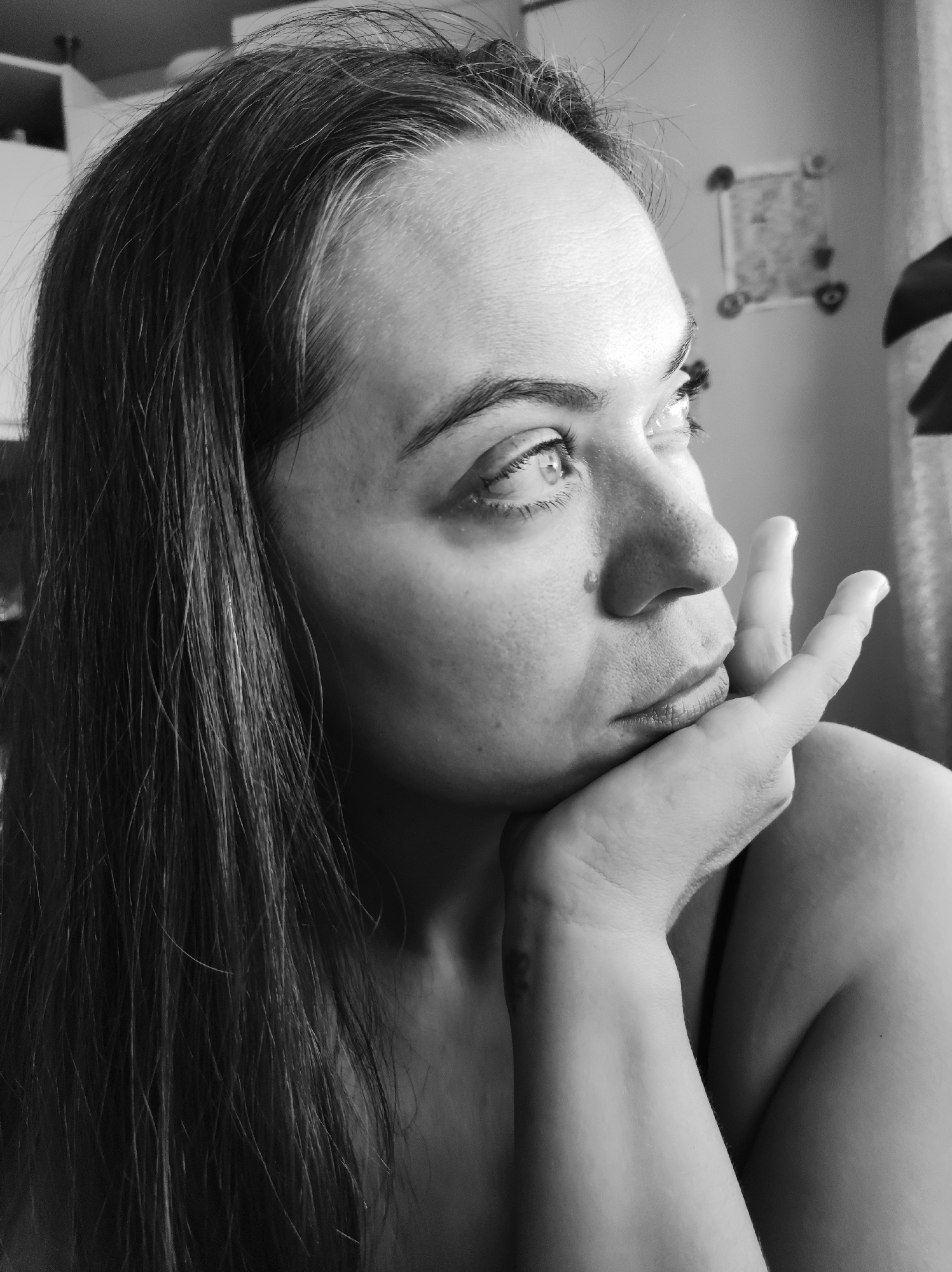 A black and white close-up photograph depicts a woman looking towards the upper right corner of the frame. The woman is positioned with her head resting on her hand, her fingers gently touching her cheek. She has long hair, with some visible grey strands, and is gazing upwards with her eyes open. The background is blurred, showing what appears to be a wall with a decorative item and a plant. The photograph is taken indoors. It is not possible to independently verify the location.