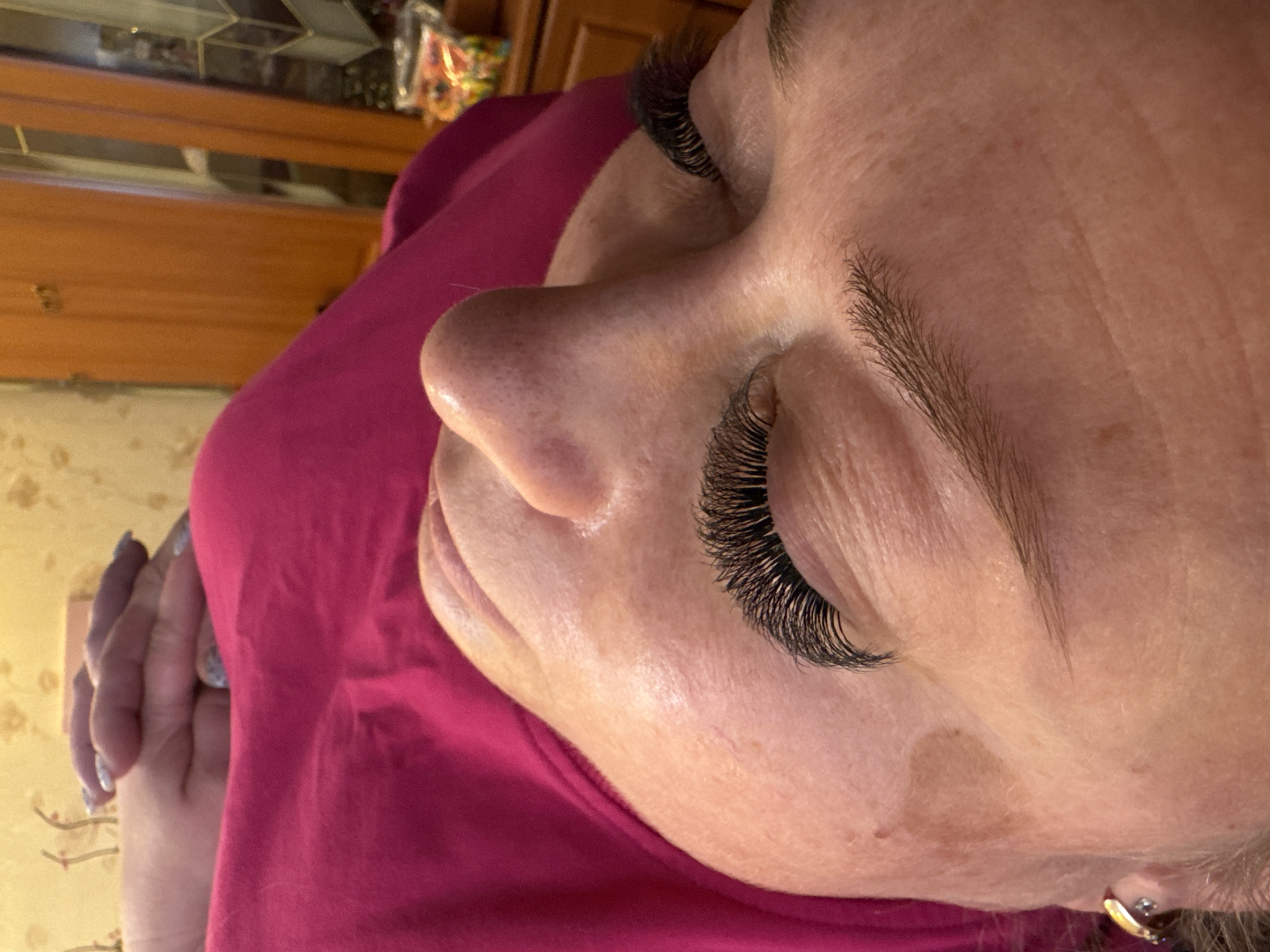 A close-up, angled photograph depicts an adult individual, likely female, lying supine with eyes closed, located in Dmitrov, Russia. The subject's head is positioned towards the left of the frame, with the body extending diagonally towards the lower right. Both eyelids feature prominent, dense, dark artificial eyelashes, curled dramatically upwards. Natural, brown eyebrows are visible above the closed eyes. The individual's skin is fair-toned, showing visible texture, some minor blemishes, and fine lines around the eyes. A gold-colored hoop earring with a small clear stone is visible in the lower left corner, affixed to the subject's left earlobe. The individual is wearing a bright magenta-colored garment, covering the upper torso. The hands are interlocked and rest on the chest area, displaying fingernails painted with a light-colored, possibly glittery, polish. The background behind the subject's head features light yellow wallpaper with a subtle patterned design. To the right, a section of a reddish-brown wooden cabinet or wardrobe is visible, and on an adjacent surface, a clear plastic bag containing various colorful objects.
