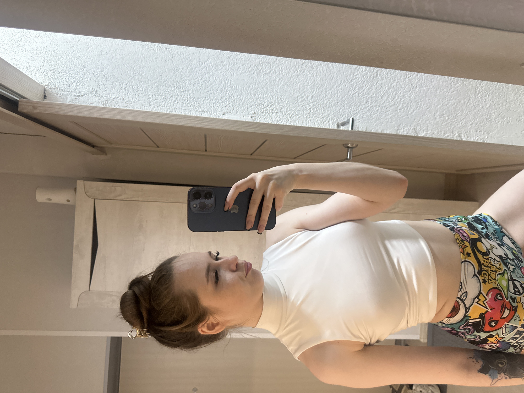 A young woman with her hair in a bun is taking a selfie in a mirror. She is wearing a white sleeveless turtleneck crop top and colorful patterned shorts. She has her left eye squinted and her mouth pursed. There is a tattoo on her left arm. The background appears to be an interior space, possibly a bedroom or dressing area, with a closet and a door visible. The lighting suggests it is daytime.