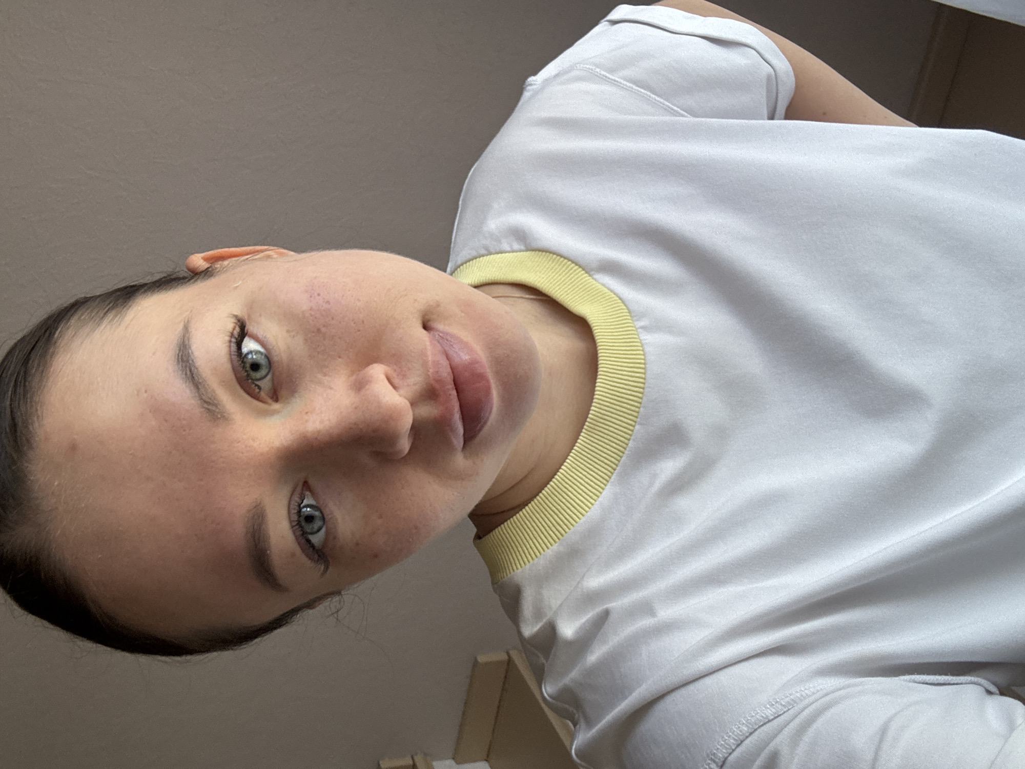 A close-up, angled shot shows a woman with fair skin, green eyes, and dark hair. She is wearing a white t-shirt with a yellow trim around the neck. The background is a light neutral color. There is no indication of the location being Ферма, Russia in the image.