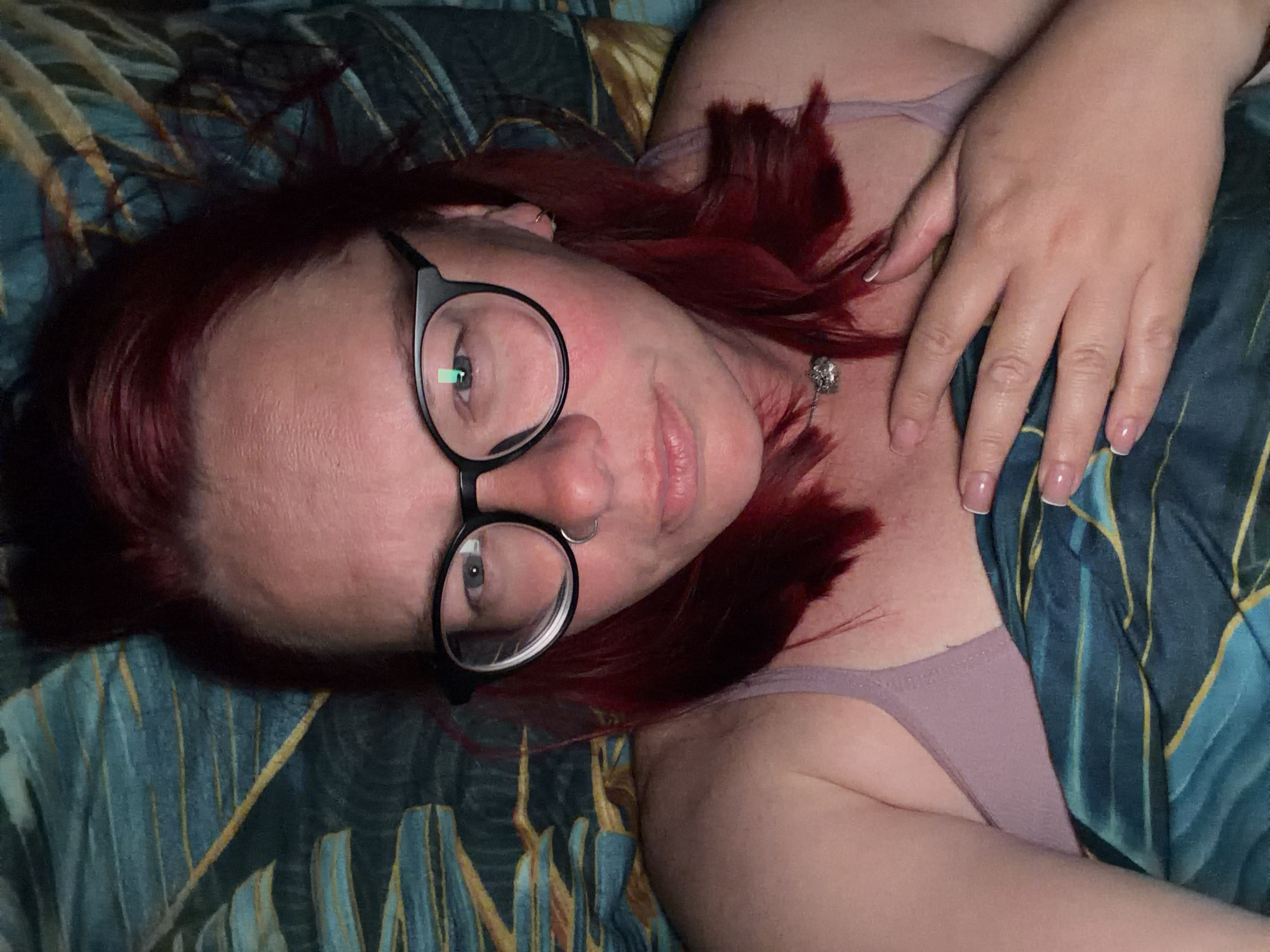A woman with red hair and glasses is lying down on a bed with a patterned blue and gold duvet cover. She is wearing a lilac-colored tank top and a necklace. Her right hand rests near her chest. The location is not evident in the image.