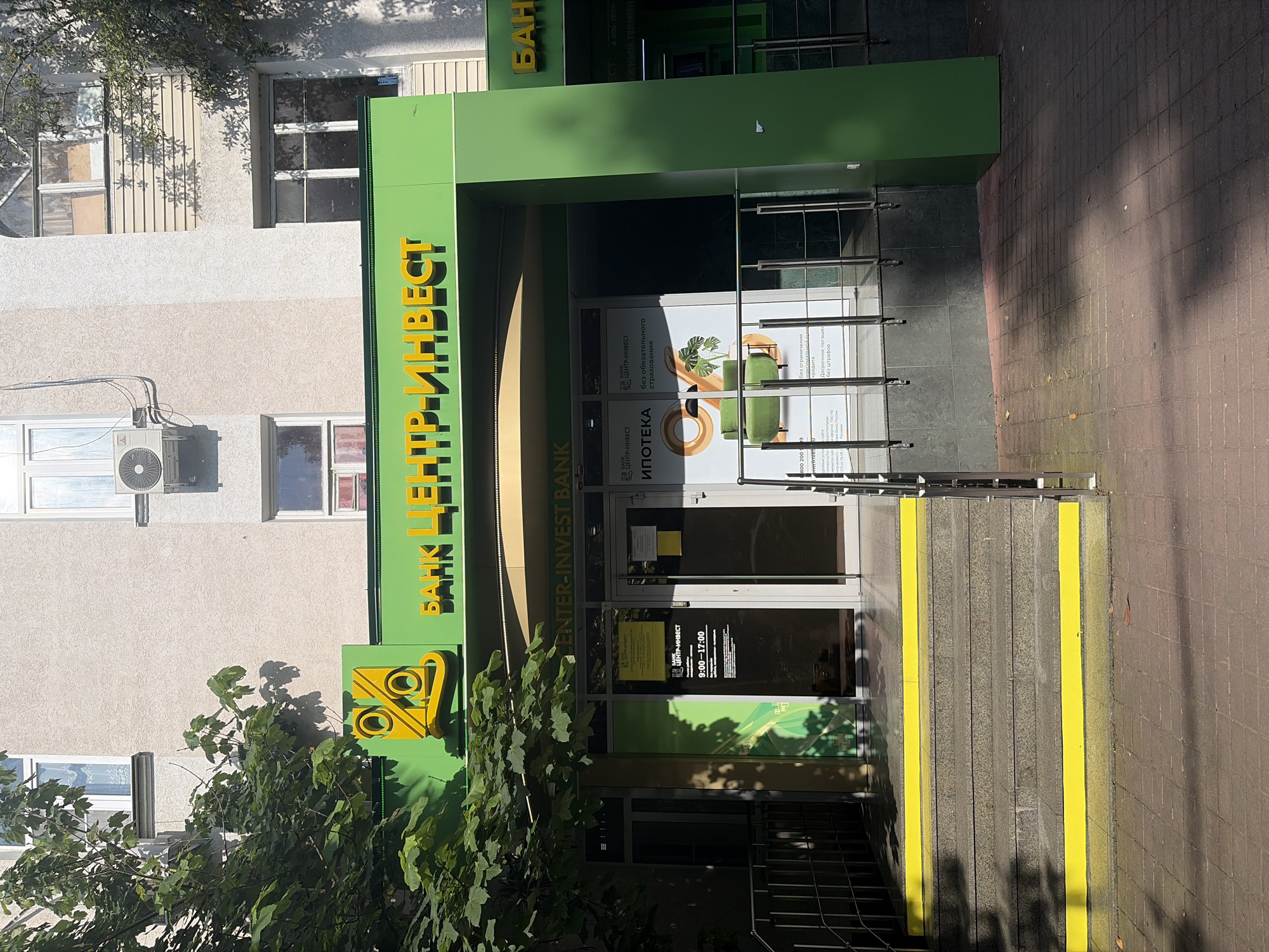 The image shows the exterior of a "Center-Invest Bank" branch in Sochi, Russia. The bank's facade is primarily green with yellow accents. A large yellow logo featuring two intertwined percentage symbols is visible above the entrance. The bank name, "BANK CENTR-INVEST," is prominently displayed in yellow lettering against the green background. The entrance features glass doors and large windows. Inside, a poster advertises "IPOTEKA" (mortgage) and "Bez obyazatel'nogo strakhovaniya kredita" (without mandatory credit insurance). The operating hours, 9:00 - 17:00, are also displayed. The building appears to be a multi-story structure with windows visible on upper floors. The entrance is accessed via a set of granite stairs with yellow safety markings on the edges. The surrounding area is paved with brick tiles, and a tree with green foliage casts shadows on the ground and the building. The lighting suggests it is daytime, likely sunny. There are no people visible in the immediate vicinity of the entrance in this shot.
