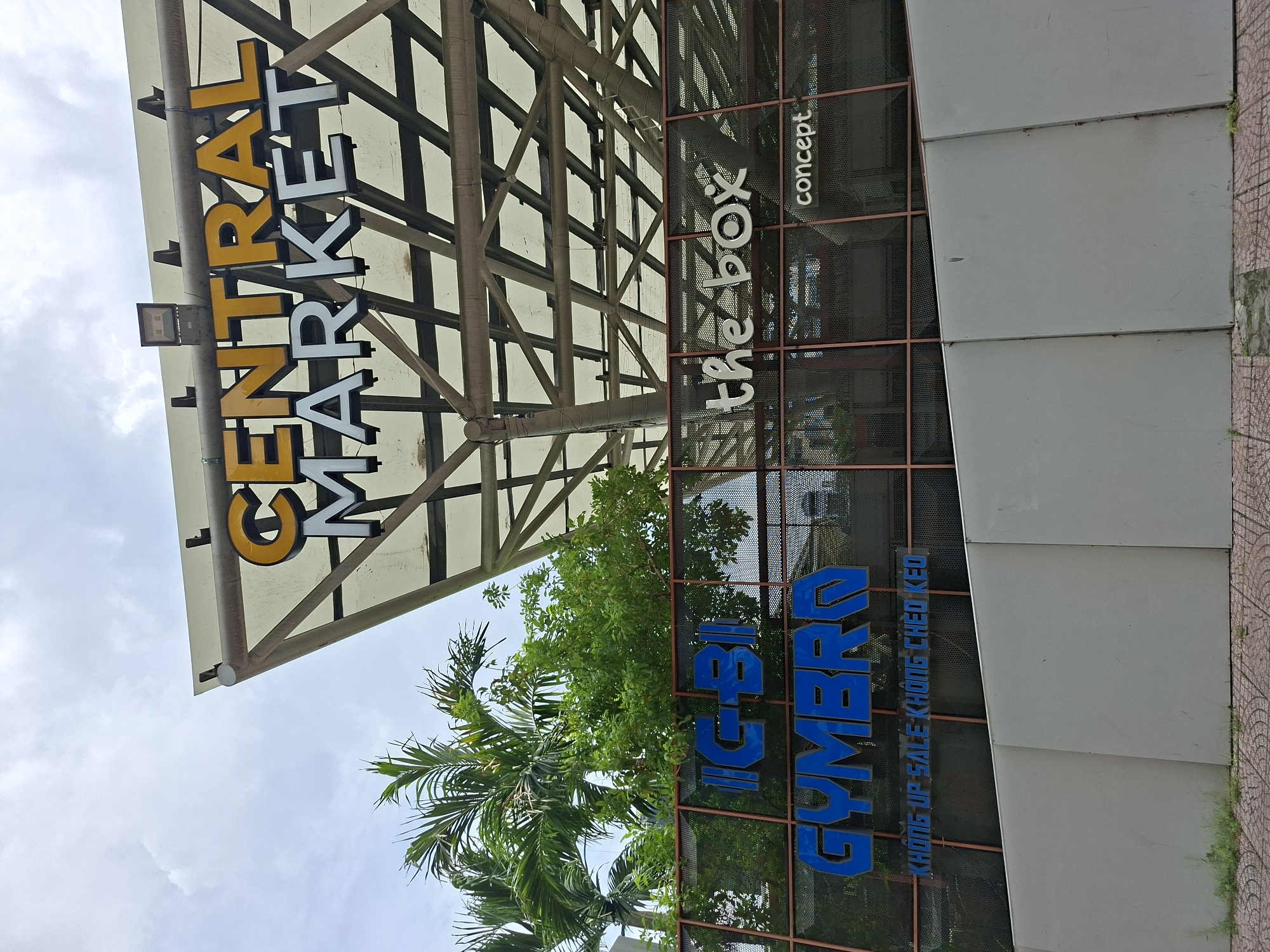 A low-angle exterior shot shows the facade of a building in Thủ Đức City, Vietnam. The building features a prominent sign with the words "CENTRAL MARKET" in large, bold lettering. The word "CENTRAL" is in yellow, and "MARKET" is in white. Below this, other signs are visible, including "IG-BI," "GYMBRO," and "the box concept." The building's architecture includes a large, angular canopy with a grid-like structure overhead. Lush green palm trees and other foliage are visible behind the signs. The ground in front of the building is paved with interlocking tiles. The sky is overcast. There are no people or animals visible in the frame.
