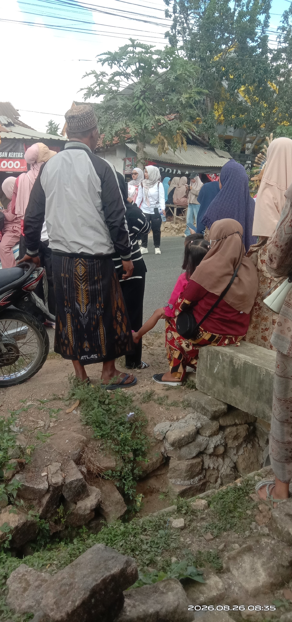 In Pakong, Indonesia, a crowd of people are gathered outdoors. In the foreground, a man wearing a traditional sarong and a jacket stands near a motorcycle. A young child in a pink shirt is being held by an adult wearing a brown hijab. In the background, other individuals in hijabs and traditional clothing are visible. The scene appears to be on a dirt path next to a road, with buildings and trees in the distance. The date and time stamp on the image indicate it was taken on August 26, 2026, at 08:35.