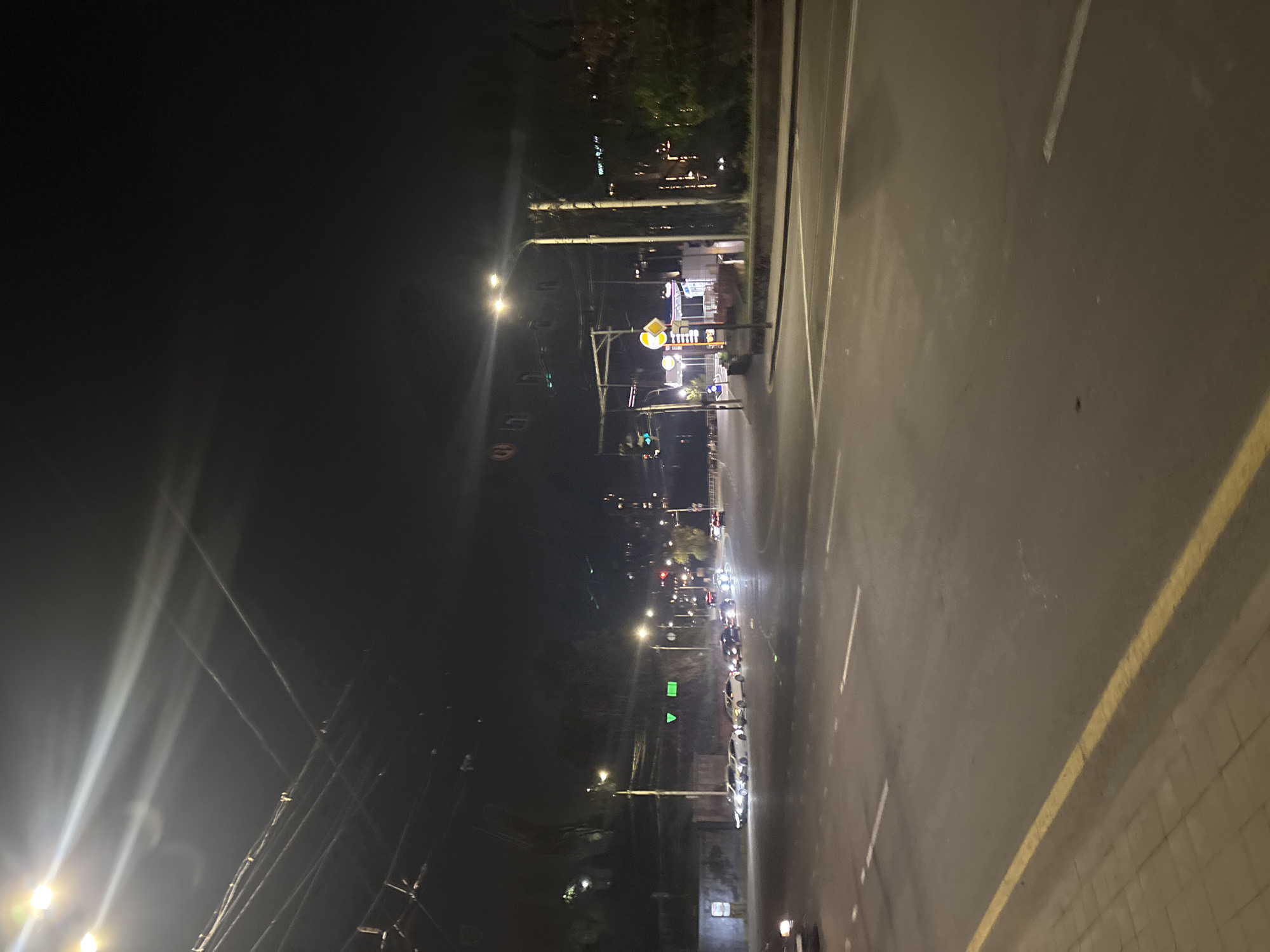 The image depicts a nighttime scene along an urban street in Yerevan, Armenia. The perspective is along the road, with a view towards a line of streetlights fading into the distance. The right side of the road has a sidewalk delineated by a yellow line. On the left side, streetlights illuminate power lines and foliage. Further down the road, cars are visible, some with their headlights on, indicating that they are in motion. In the middle distance, a store or restaurant is lit up, with street signs visible in front.