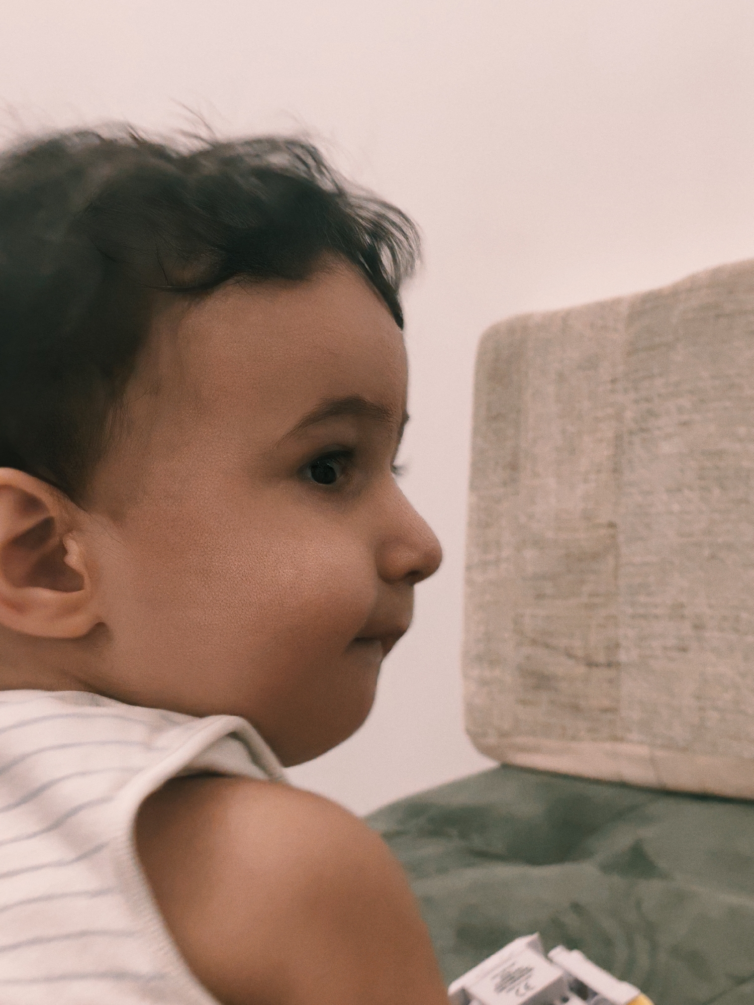A close-up, profile shot captures a young child's face, seen from the left side, against a pale, out-of-focus background. The child has dark, wispy hair, a visible ear, and their eye and nose are in focus. They are wearing a white tank top with thin gray stripes. In the lower right corner of the image, a portion of a white toy with yellow accents is visible, resting on a green surface. The background consists of a textured, light-colored object on the upper right and a soft, green surface below it. The lighting is soft, creating a slightly blurred effect overall, with the exception of the child's facial features.