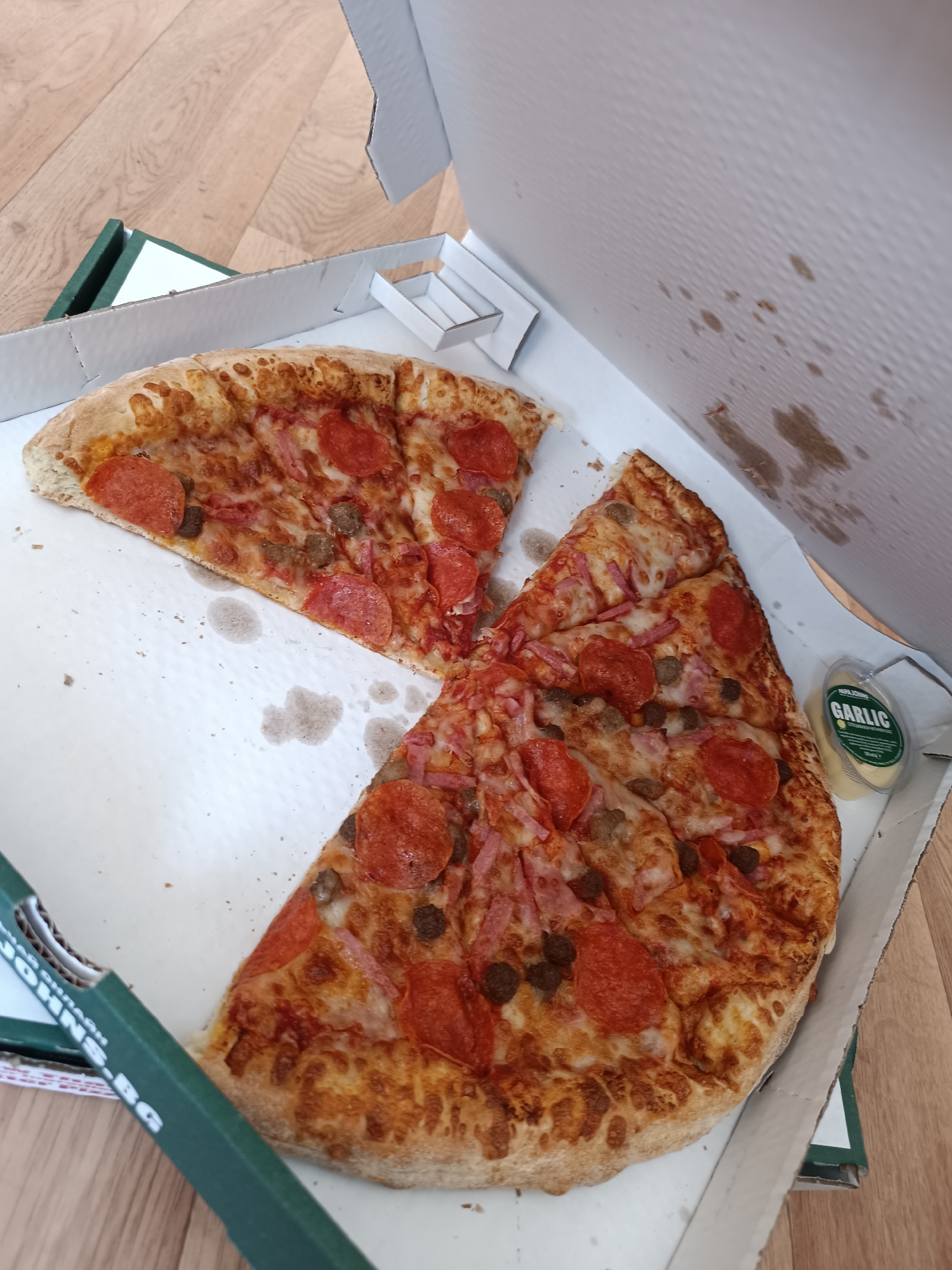 The image shows an open pizza box containing two slices of pizza with pepperoni, ham, and sausage toppings, along with a small container of garlic sauce. The box appears to be from Papa John's, based on the visible text "JOHNS.BG" on the side of the box. Given the ".BG" extension, it is likely that the location is in Sofia, Bulgaria. The pizza box is resting on a wooden floor. The interior of the pizza box has grease stains on the sides.