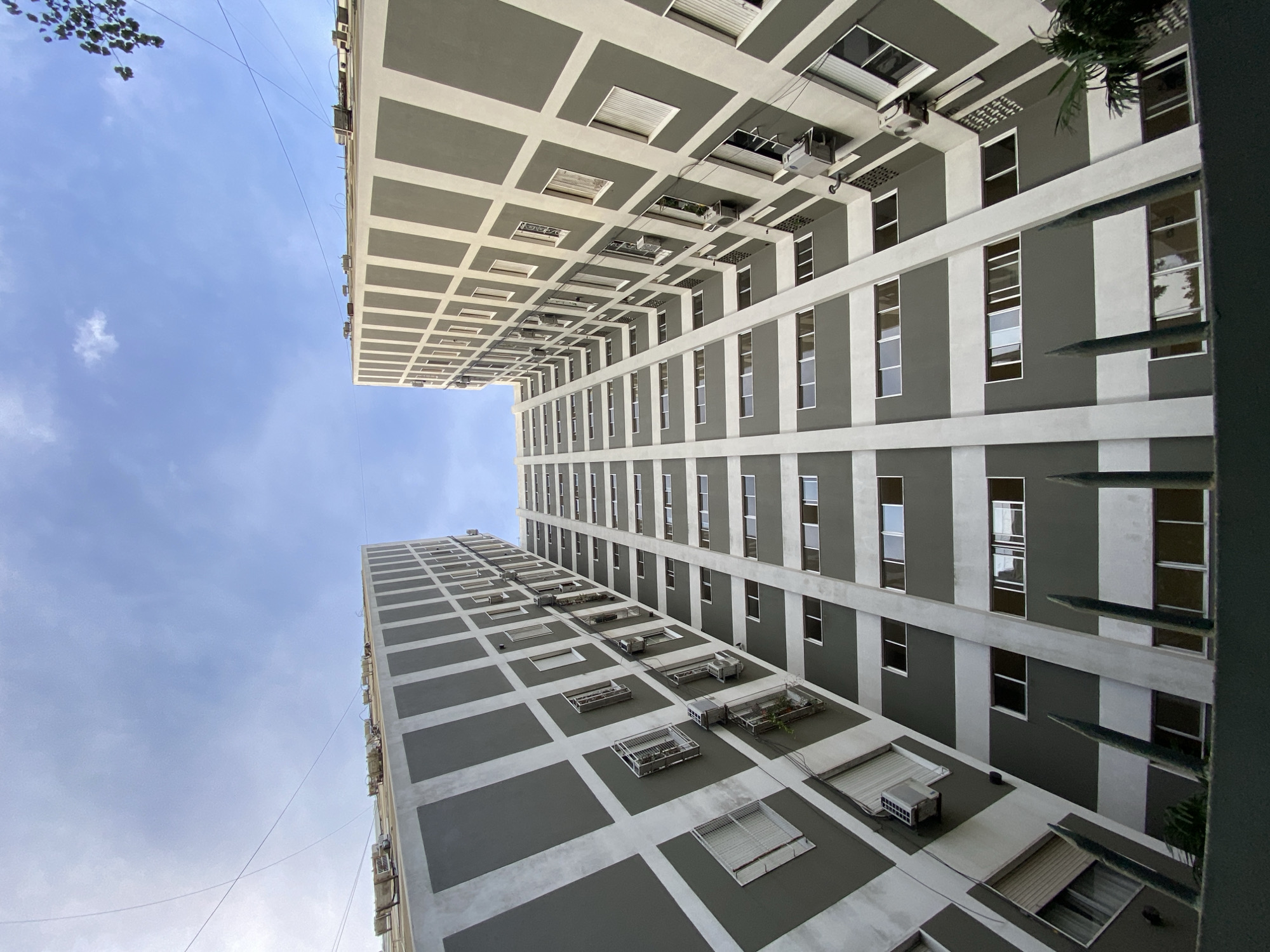 The image is a low-angle shot of two tall buildings in Buenos Aires, Argentina. The buildings are similar in design, with a gray and white color scheme. They have a rectangular shape with windows arranged in vertical rows. There are air conditioning units visible on the exterior of the buildings. The sky is visible between the buildings, with some clouds present. There are a few tree branches visible in the top left corner.