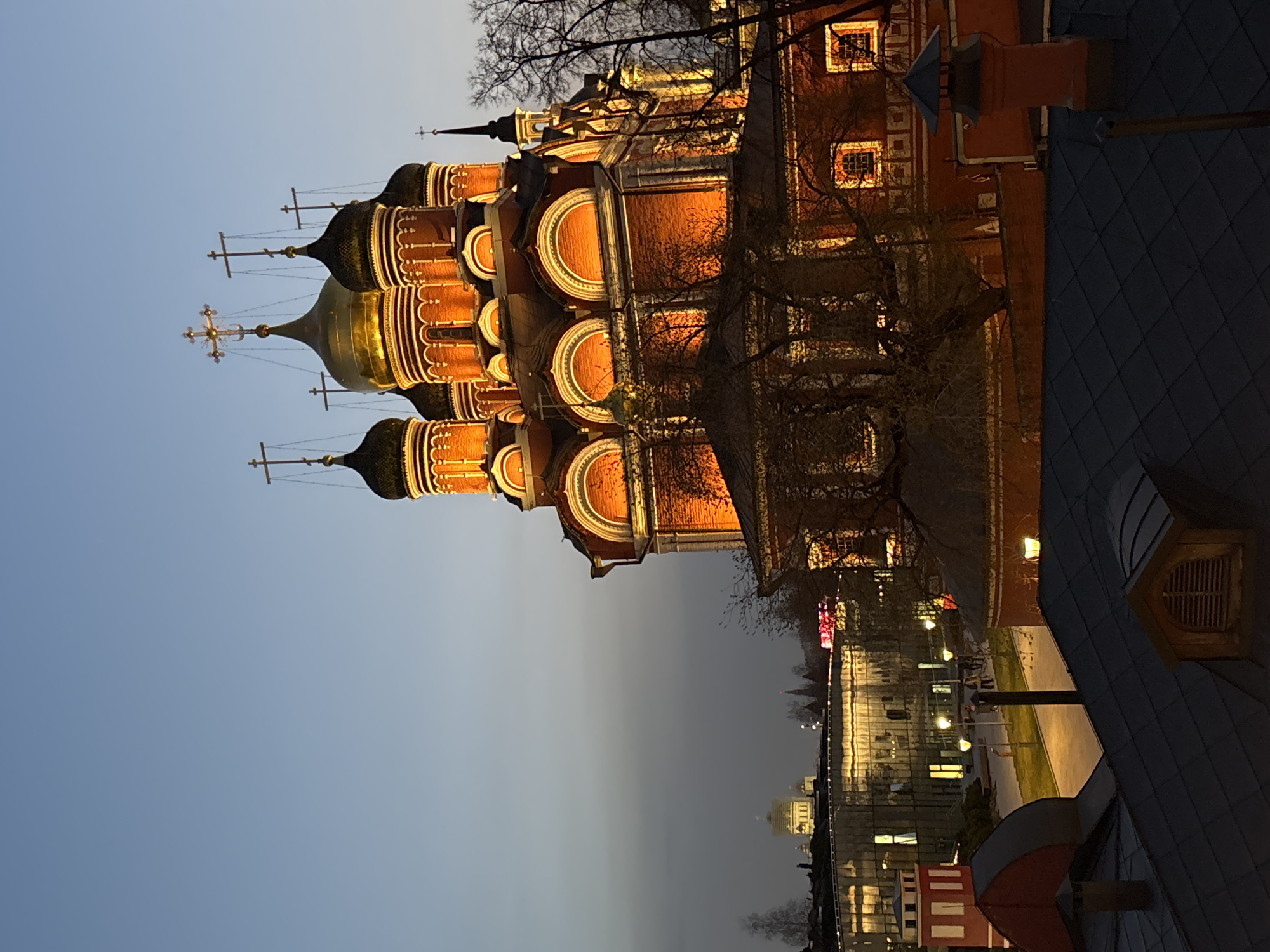 The image shows a view of a Russian Orthodox church with multiple onion domes, illuminated by warm light against a twilight sky. The church's architecture includes brick facades and arched decorative elements. In the background, city buildings and lights are visible. The image is taken from a high vantage point, possibly a rooftop, with details of the rooftop visible in the foreground. The location is likely in Russia, given the architectural style.