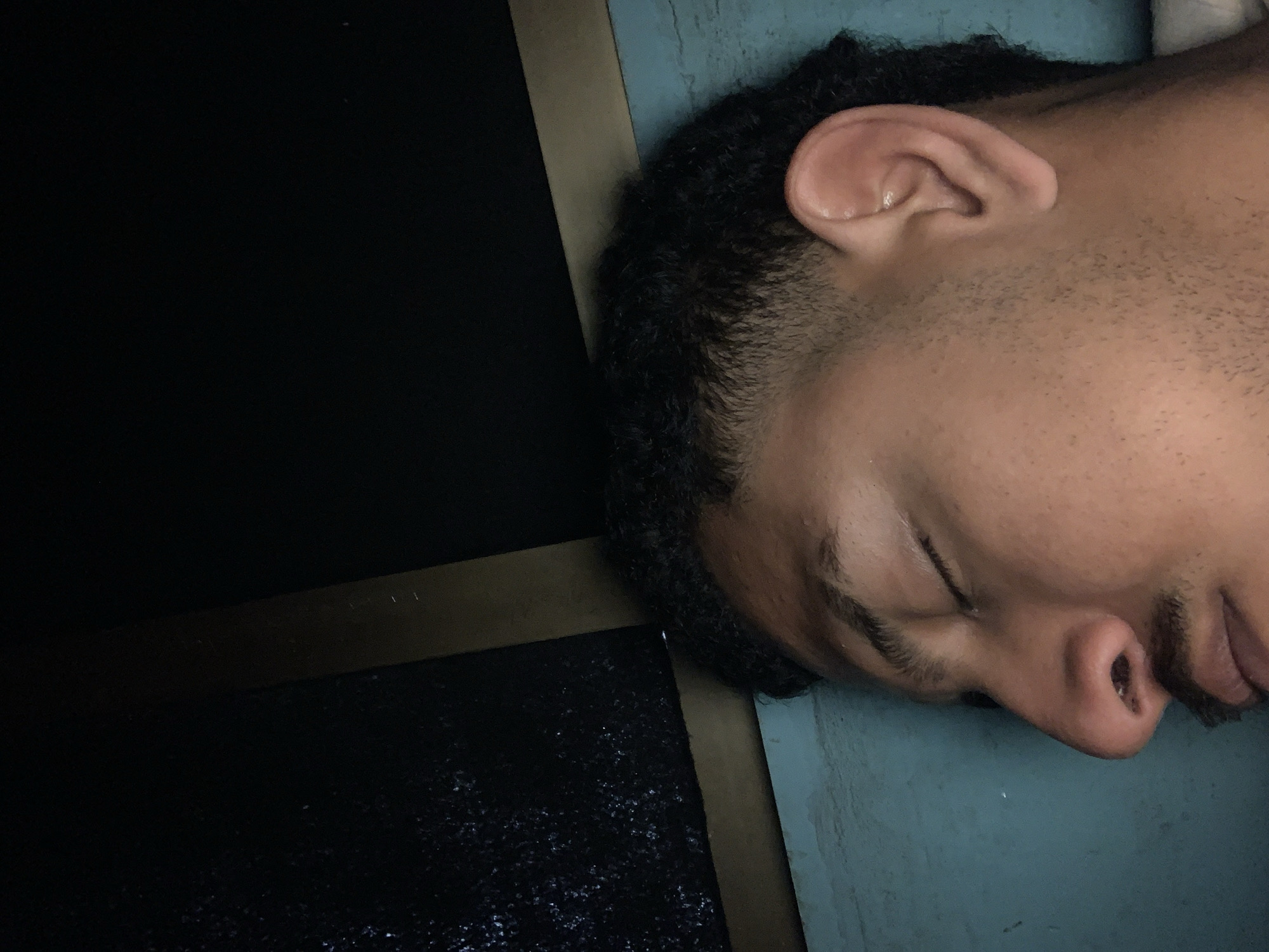 The image shows a close-up, side profile of a person's face, with their eyes closed, appearing to be asleep. The person has dark, curly hair, a visible ear, and some facial hair. The background is dark, with a textured area on the left and a wooden beam or edge running horizontally across the upper portion of the frame. The person is lying on a surface that appears to be a turquoise-colored wall or furniture. The scene is indoors, with low lighting. There is no specific information to indicate this location is in Santa Ana, Venezuela.