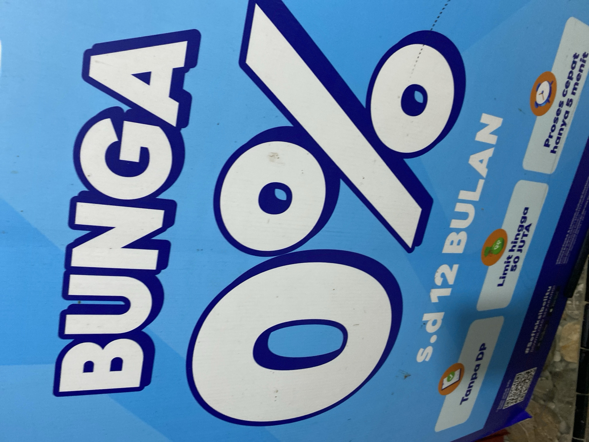 The image shows a close-up of a blue banner with large white text that reads "BUNGA 0%". Below this, text reads "S.d 12 BULAN". There are also three smaller white rectangles with text and icons. The first rectangle on the left says "Tanpa DP" with an icon of a phone and a checkmark. The second rectangle in the middle says "Limit hingga 50 JUTA" with a money bag icon. The third rectangle on the right says "Proses cepat hanya 5 menit" with a clock icon. At the bottom left of the image, a QR code is visible, and the hashtag "#Softekbelitu" is written in white text. The banner appears to be displayed outdoors, possibly on a wall or a street. There are no people visible in the image. The lighting suggests it is daytime. This banner is likely promoting a financial product or service, possibly a loan or credit offer, with 0% interest for up to 12 months, no down payment required, a limit of up to 50 million (JUTA), and a fast processing time of 5 minutes. The mention of "Ta', Indonesia" in the prompt is not visually verifiable from the provided image itself.
