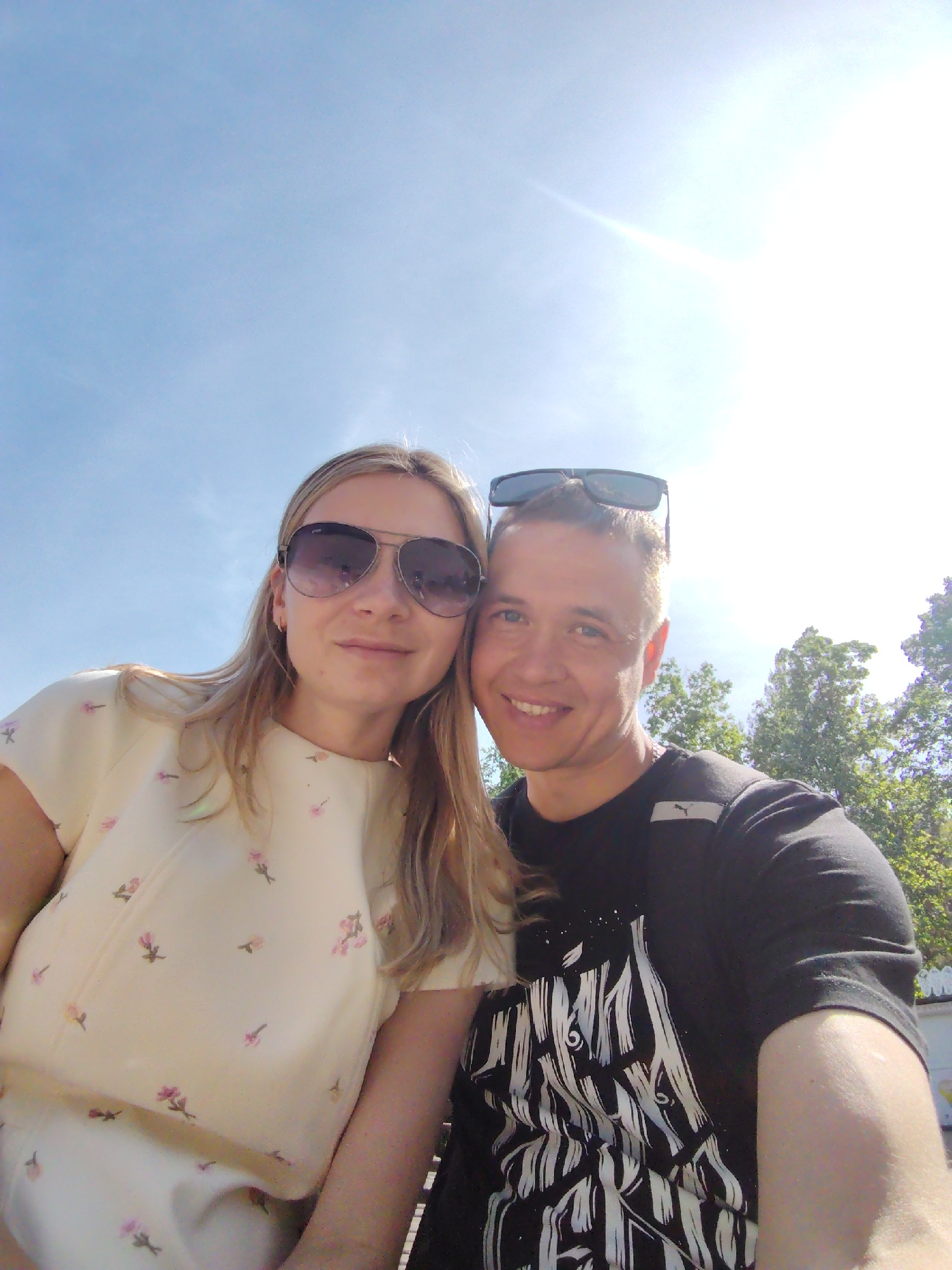 A selfie captures a couple in Chaikovsky, Russia. The woman wears sunglasses and a dress with floral patterns. The man has sunglasses perched on his head and a black t-shirt with a graphic design. They are outside on a sunny day, with a blue sky above and trees in the background.