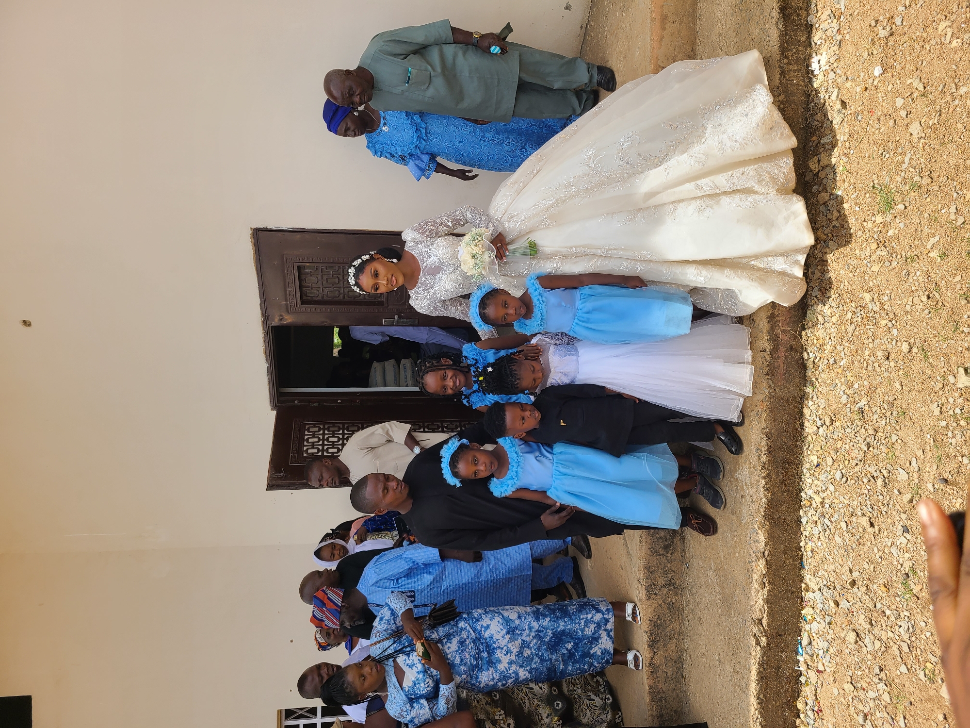 A wedding party poses for a photograph outside a building in Barakin Lerawi, Nigeria. The bride, wearing a white gown with a floral bouquet, stands in the center. Several flower girls wearing light blue dresses are positioned at the front and beside the bride. Two older men and a woman in a blue dress stand behind the bride. Other wedding guests are gathered on the left side of the scene. The location has beige walls and the ground is a mix of dirt and small rocks.