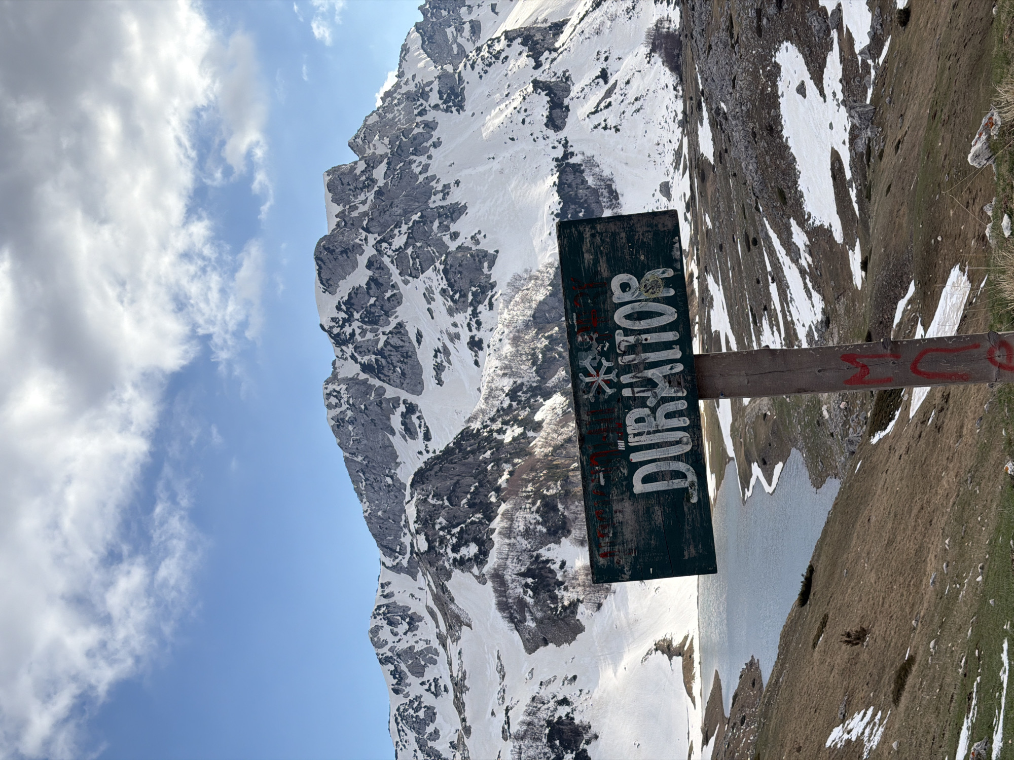 The image shows a landscape in Pašina Voda, Montenegro. A sign with "DURMITOR" written vertically is in the foreground. In the background, a snow-covered mountain rises against a partly cloudy sky. A body of water is also visible, partially obscured by the terrain. The foreground consists of grass and some snow patches.