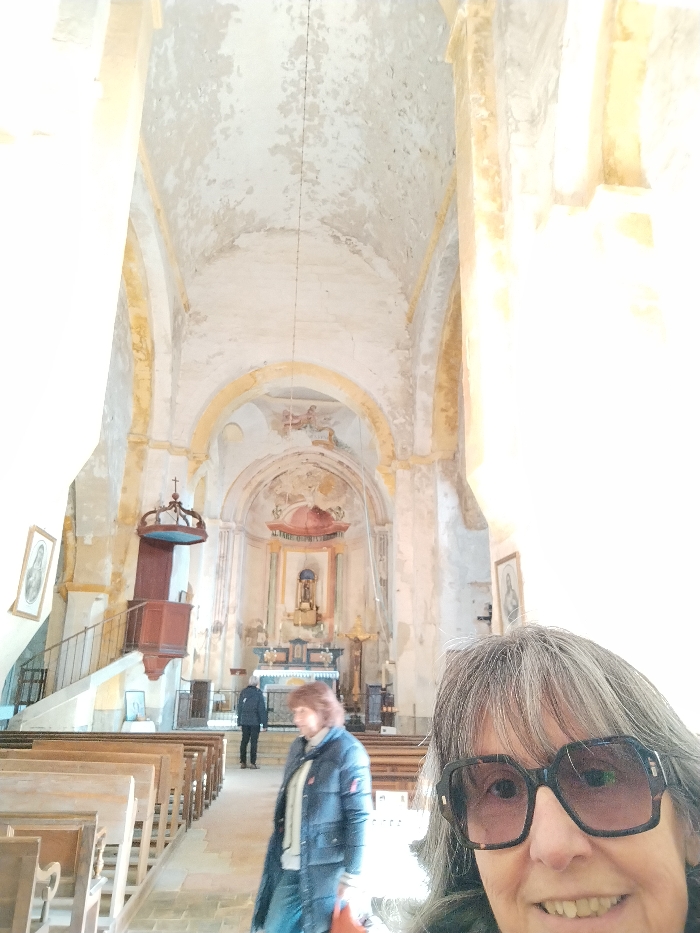 An interior shot of a church or cathedral, seemingly taken as a selfie. A woman with grey hair and large glasses smiles into the camera. Behind her, two other people are seen walking towards the altar. The interior features high ceilings with faded paintings, arches, a wooden pulpit, and rows of pews. The walls are aged, with peeling paint, suggesting a building of historical significance.