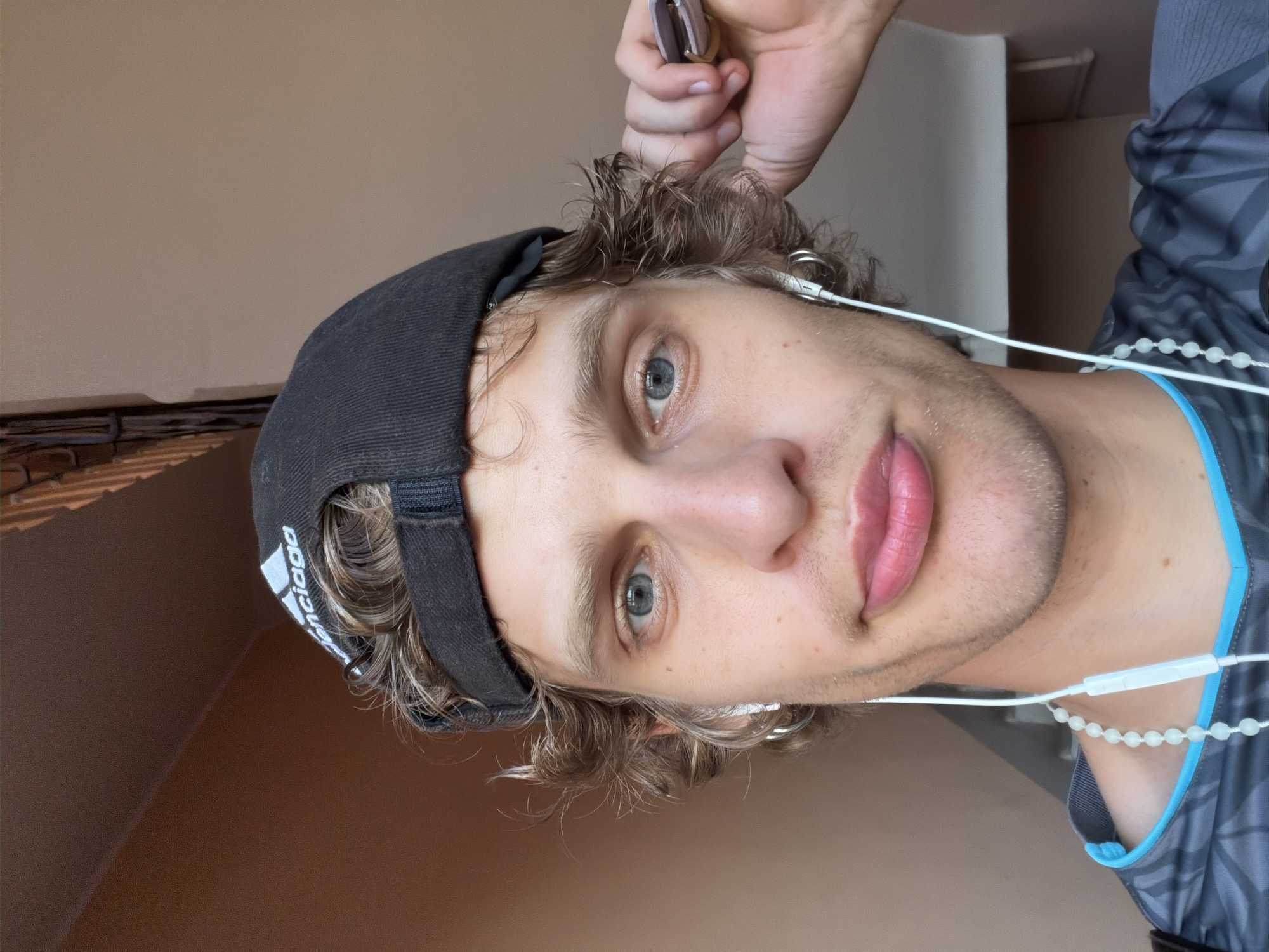 A person is depicted in a close-up, angled shot. The individual has curly, light brown hair and light-colored eyes. They are wearing a black baseball cap with white text. The person has earbuds in their ears and is wearing a gray and blue shirt. The background appears to be an interior setting with a wall. There is no information in the image to determine the location is Milan, Italy.