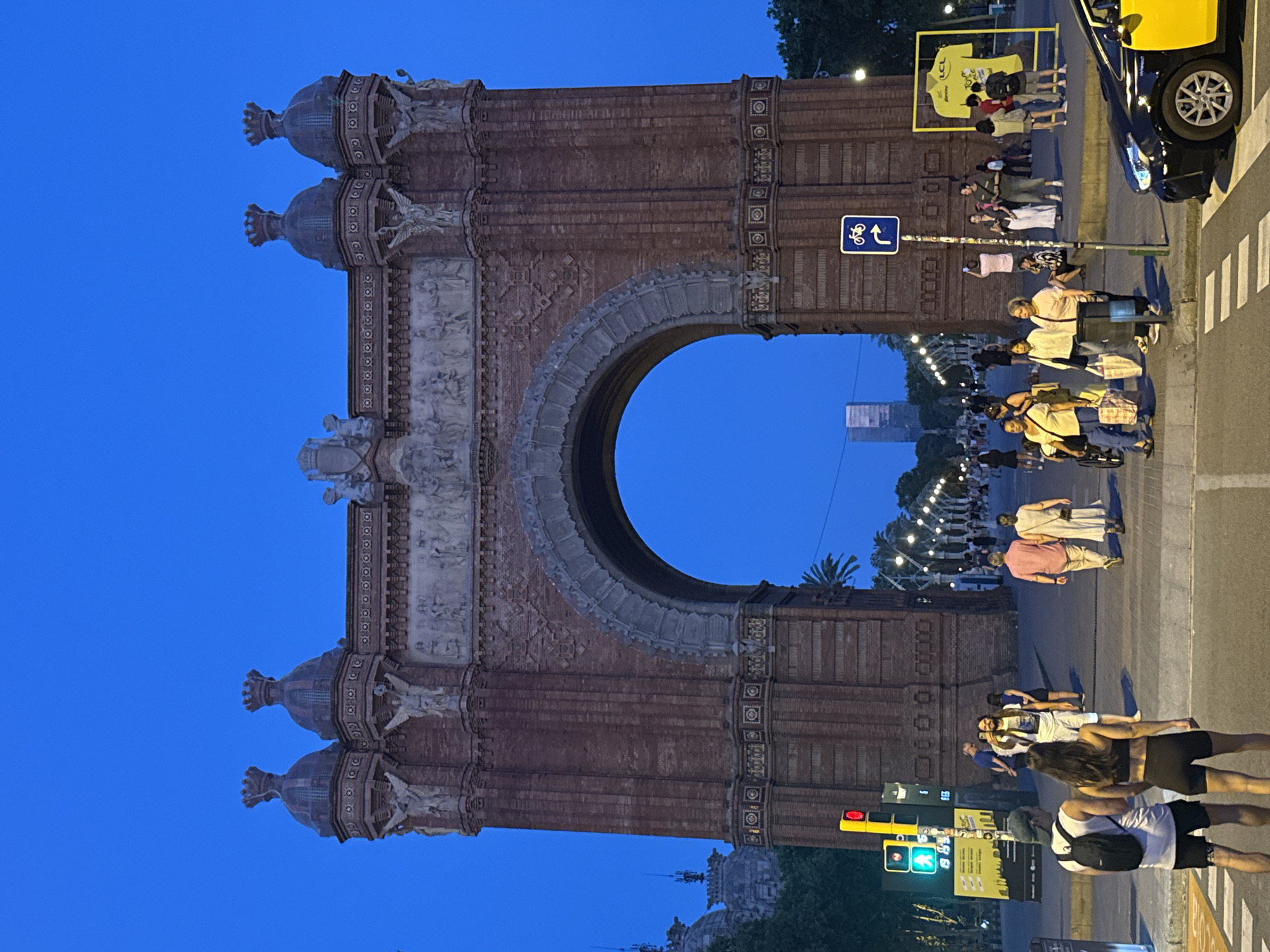 In Barcelona, Spain, the Arc de Triomf stands as a grand monument under a deep blue twilight sky. The arch, constructed of red brick, is adorned with intricate sculptures and topped with ornate finials. A wide avenue stretches out behind it, lined with lampposts and palm trees, leading towards a distant skyscraper. A diverse group of pedestrians is gathered near the arch, some walking, others taking photographs, and a few are conversing. A black and yellow taxi is parked in the foreground, facing away from the camera, on a street with pedestrian crossings. Traffic lights are visible, indicating a busy urban environment. The scene captures a lively evening atmosphere at a prominent landmark.