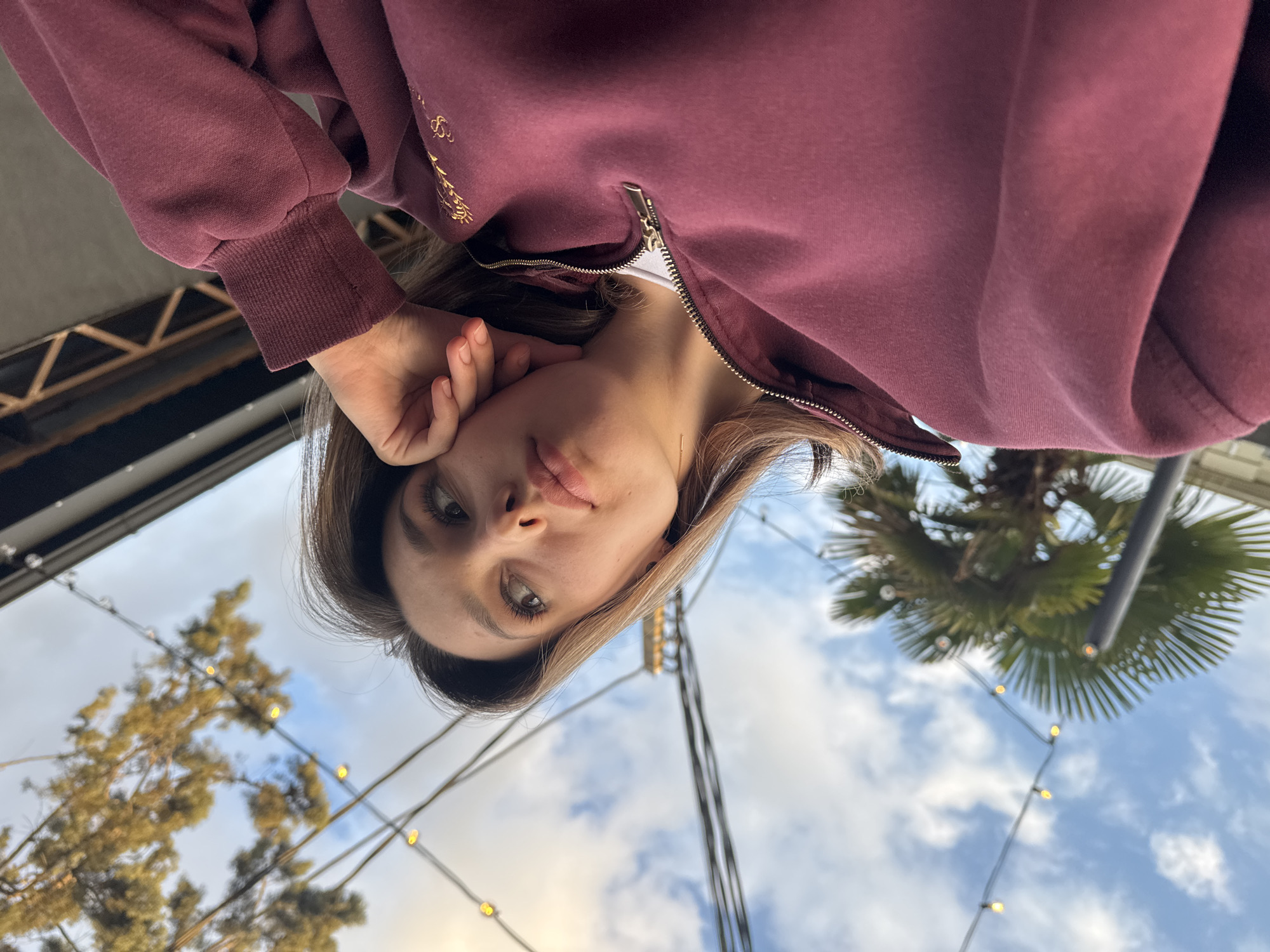 A low-angle, head-and-shoulders portrait of a young woman looks directly at the camera from below. The image appears to be taken from a perspective looking down at her, with the background seen above her head. She has light brown hair, which is parted on her left, framing her face. Her eyes are a light color, possibly green or hazel, and she has a slightly pouting or contemplative expression. Her left hand is raised, with fingers gently touching her chin or cheek. She is wearing a maroon or dark reddish-brown zip-up hooded sweatshirt. On the right side of her chest (from the viewer's perspective, her left), there is gold-colored embroidery, possibly stylized text or a logo, though it is not clearly legible. A thin gold chain necklace with a small pendant is also visible around her neck.
The background is an outdoor setting under a partly cloudy blue sky. Strands of decorative string lights with small, glowing yellow bulbs are strung across the frame, creating a festive atmosphere. To the left, a deciduous tree with light-colored branches is visible, while to the right, a palm tree with green fronds stands out against the sky. In the upper left corner of the frame, dark, possibly metal, architectural elements like beams or rafters suggest an overhead structure such as a pergola or awning. The lighting is soft and natural, indicating daylight, possibly an overcast day or late afternoon, given the visible clouds and the ambient light. There is no clearly readable text in the image.