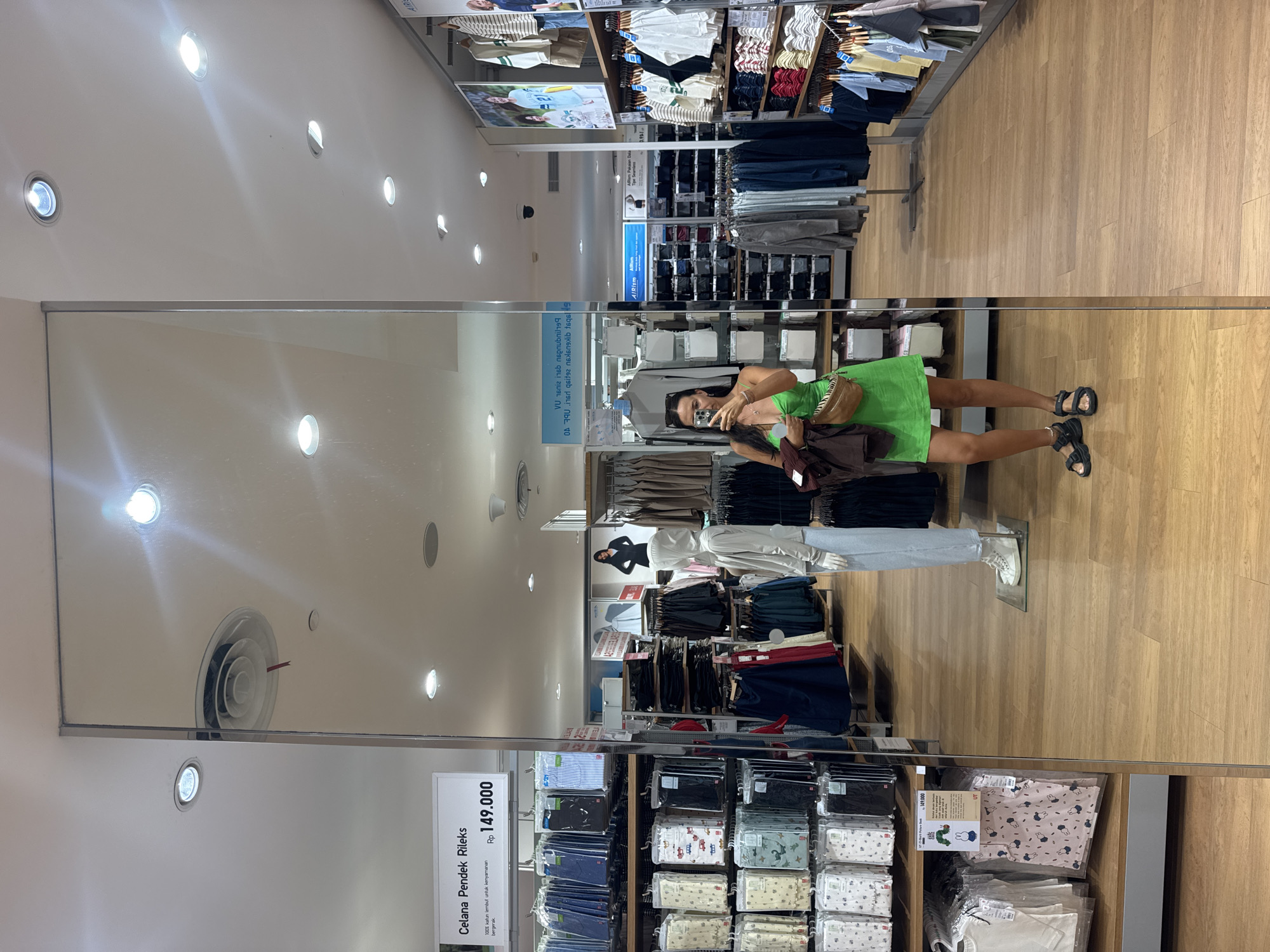 A woman is taking a mirror selfie in a retail store, identified as being in Kuta, Indonesia. The store appears to be a clothing store, likely Uniqlo, based on the signage and merchandise displayed. The woman is wearing a bright green mini dress, black platform sandals, and a brown fanny pack. She is holding a dark brown jacket in her left arm. The mirror reflects the store's interior, showing racks of clothing, mannequins, and store displays. The floor is made of light-colored wood. The lighting in the store is bright, with overhead lights visible. The reflection in the mirror also shows ceiling features such as air vents and recessed lighting.