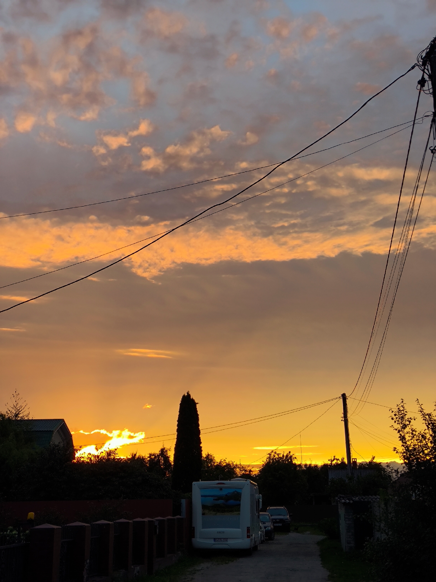 An exterior, eye-level, vertical shot captures a vibrant sunset over a rural or suburban landscape. The sky is dominated by hues of orange and yellow, with scattered clouds tinged with pink and grey. Power lines crisscross the upper portion of the frame, silhouetted against the colorful sky.
In the foreground, a white camper van is parked on a dirt road, facing away from the camera. To the left of the van, a dark fence runs horizontally across the lower part of the image. Behind the fence, dense green foliage and trees are visible. A tall, slender tree stands prominently in the middle ground, to the left of the camper van, and a house with a green roof is partially obscured by trees.
To the right of the camper van, two cars are parked on the road, also facing away from the camera. Further back, a utility pole with multiple wires stands on the right side of the road. The overall scene suggests a tranquil evening setting in a location that could be on the outskirts of Kaliningrad, Russia, characterized by its combination of residential structures, natural elements, and infrastructure.