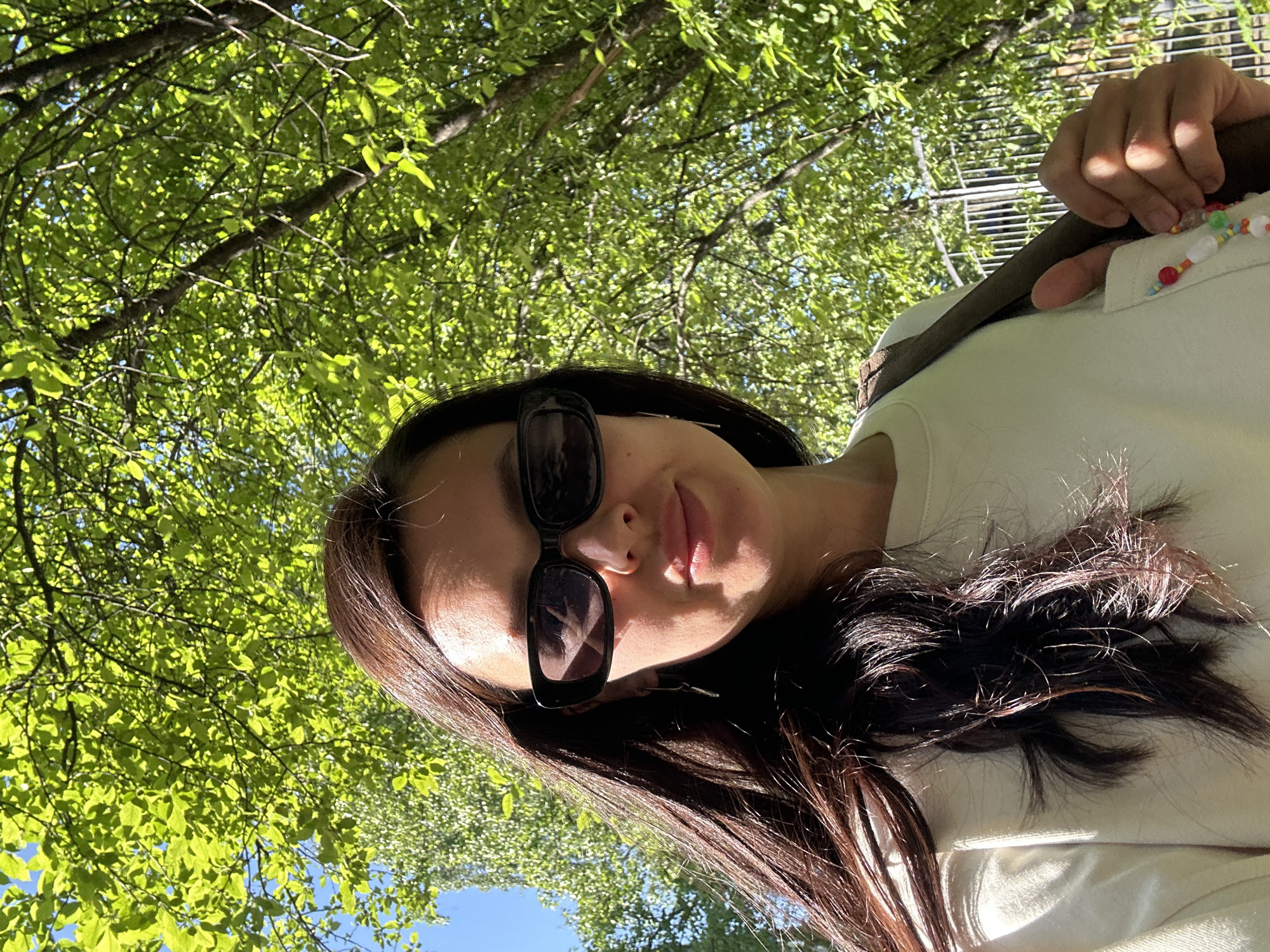 A woman wearing sunglasses and a light-colored shirt with a beaded strap is photographed from a low angle. The background consists of green foliage, suggesting a wooded area. The presence of the trees suggests the location could be a park or green space within the city of Chaikovsky, Russia.