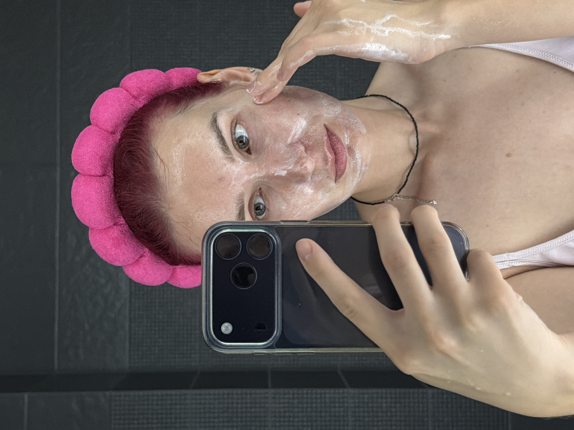 A young adult with pink hair and a bright pink fluffy headband is taking a selfie in a bathroom. They are applying a white cream or cleanser to their face with their right hand while holding a black smartphone in their left hand, with the camera facing them. The background appears to be a dark, tiled wall. The individual is wearing a light pink camisole and a thin black necklace with a small silver charm. The image is cropped to focus on the person's face and upper body. This image was taken in Chaikovsky, Russia.