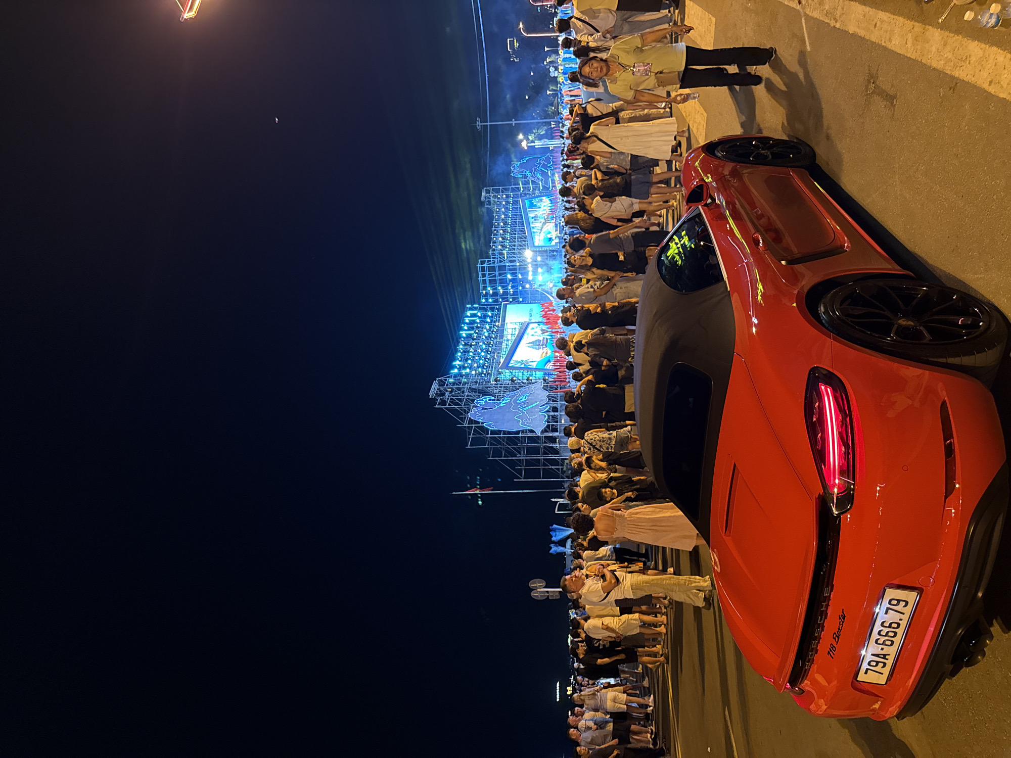 This is a photograph taken at night in Nha Trang, Vietnam, likely during an outdoor event or festival. In the foreground, a red convertible Porsche 718 Boxster is parked on an asphalt road with yellow lane markings. The car's convertible top is down. The license plate reads "79A-666.79". Behind the car, a large crowd of people is gathered, facing a brightly lit stage or elaborate light installation in the background. The stage appears to be a temporary structure made of scaffolding, adorned with numerous lights and large screens displaying images. The sky is dark, suggesting it is nighttime. The environment appears to be an open area, possibly a waterfront promenade or public square, with streetlights visible in the distance. The people in the crowd are dressed in casual summer attire. A woman in a light-colored dress is standing near the front of the crowd, and a man in a light-colored shirt and pants is also prominent. The overall atmosphere suggests a lively event.