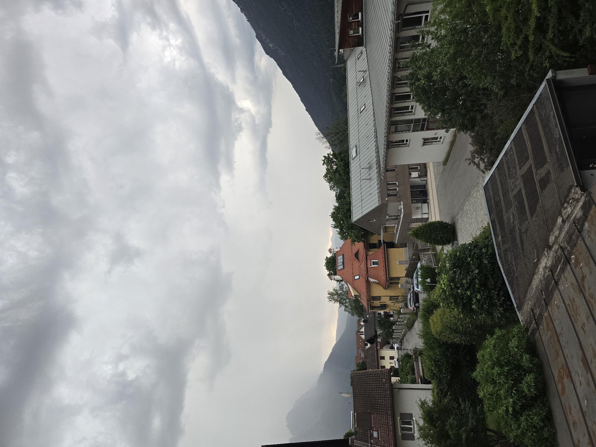 The image shows a view of a town with mountains in the background under a cloudy sky. The town appears to be located in a valley, with buildings nestled among trees. The architecture suggests a European setting, possibly in the Alps. The lighting indicates it might be overcast or nearing dusk, with a slight haze over the distant mountains. There are no people visible in the image. The foreground shows the roofs of buildings, one of which appears to be made of corrugated metal. The middle ground features houses with pitched roofs, some with vibrant colors, and a larger, more modern building with a flat, metal roof and many windows. The background is dominated by the imposing presence of the mountains, partly obscured by the weather. The overall impression is of a quiet, residential area in a mountainous region.