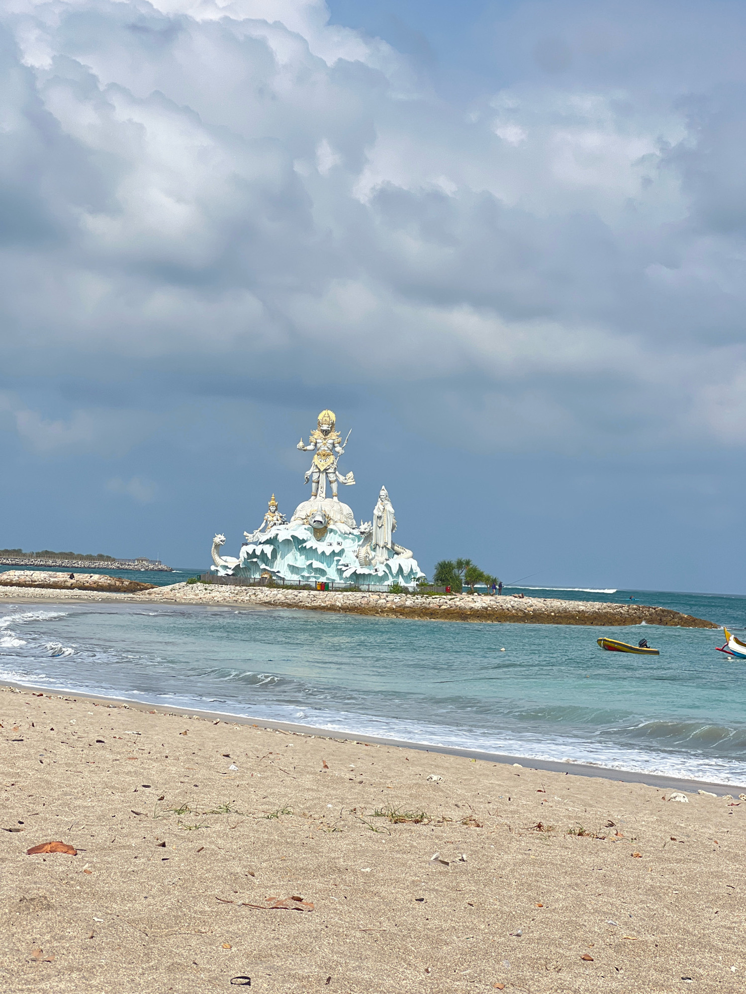 A wide-angle, eye-level shot captures the Dewa Ruci statue, a prominent landmark in Kuta, Bali, Indonesia, standing majestically on a rocky island in the ocean. The statue, a depiction of the Hindu deity Dewa Ruci, is a monumental white and gold structure situated atop a blue and white wave-like base. Flanking the central figure are two smaller statues, one on the left with a dragon-like creature and another on the right. The statue is surrounded by a rocky breakwater, with lush green vegetation visible on the island.
The foreground is dominated by a sandy beach, with small pieces of debris scattered across its surface. Gentle waves with white foam lap at the shore, leading to the turquoise ocean. Several small, colorful boats are anchored in the water, with one yellow and red boat prominently featured to the right. A causeway, possibly a pier, extends from the mainland towards the island. The sky above is filled with dramatic, gray clouds, suggesting an overcast day, yet the sunlight manages to illuminate the scene, casting a soft light. The overall atmosphere is serene and draws attention to the cultural and natural beauty of Kuta.