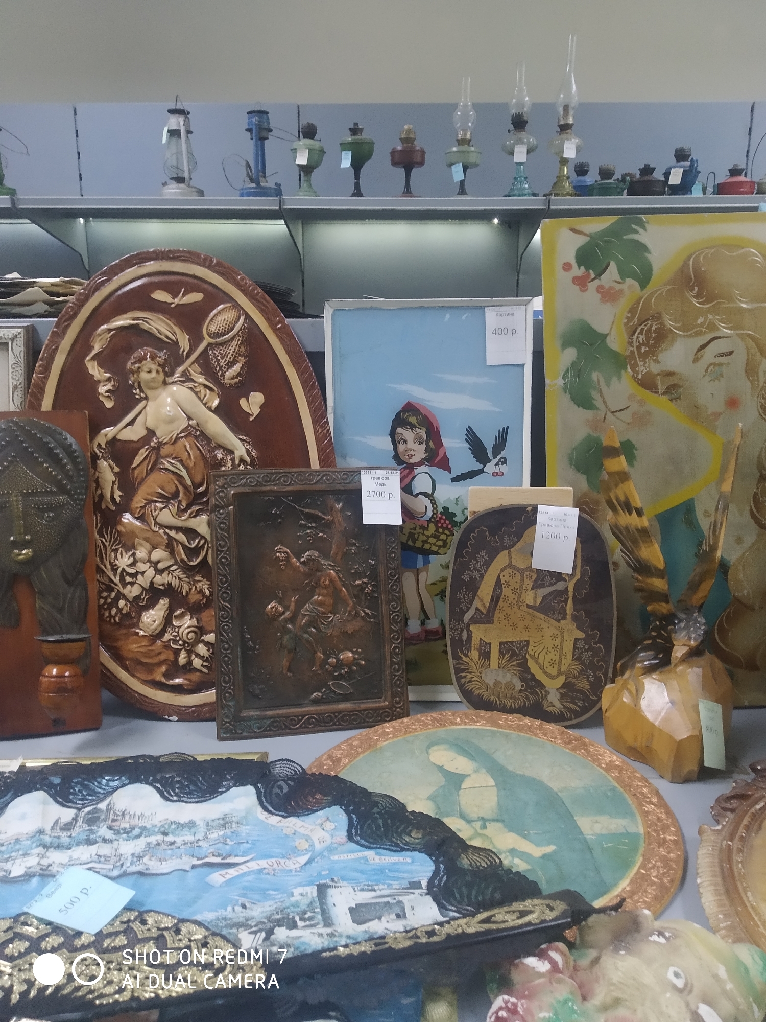 The image shows a display of various decorative items and artwork, likely for sale, within a store in the city of Chaikovsky, Russia. The items are arranged on a shelf against a neutral background.
The items include:
- A large, oval, brown plaque with a relief of a woman with a net, possibly representing a mythological figure.
- A framed picture of a cartoon girl with a red hood, holding a basket, with a bird flying nearby. A price tag indicates a price of 400 rubles.
- A framed copper relief depicting figures in a woodland scene; a price tag shows 2700 rubles.
- An oval plaque with a golden image of a woman on a swing; a price tag shows 1200 rubles.
- An oval plaque featuring the Madonna and Child in muted blue and green tones.
- A souvenir picture of "Mallorca" in a black frame with a lace trim; a price tag shows 500 rubles.
- A carved wooden figure with feathers.
- Several antique oil lamps are on the shelf above.
The items appear to be a mix of vintage and newer decorative pieces. The lighting is somewhat diffused, and the objects are closely packed together. Text at the bottom indicates the image was taken with a Redmi 7 AI Dual Camera.