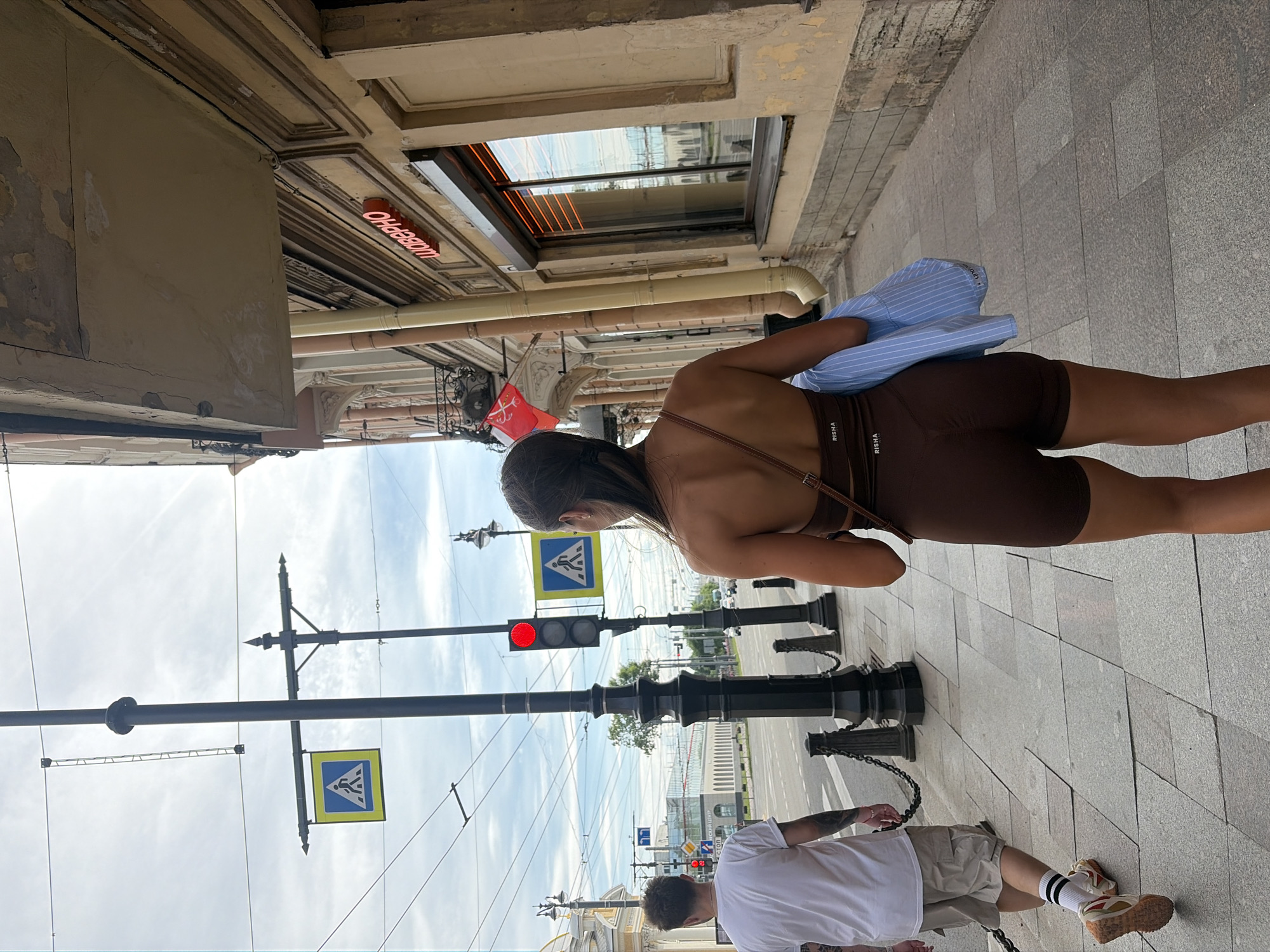 An urban street scene in Saint Petersburg, Russia, is depicted under a partly cloudy sky. Two individuals are walking on a grey paved sidewalk.
In the foreground, a female subject is viewed from behind, walking towards the viewer's right. She has dark hair tied back and wears a dark brown, form-fitting athletic top and matching shorts, both with "RISHA" printed in white text. A light blue striped shirt is draped over her left arm. A thin brown strap, likely from a bag, extends over her right shoulder.
Ahead and to the left of the female, a male subject is also walking away from the viewer. He has short dark hair and tattoos on his left arm. He wears a white t-shirt, light khaki cargo shorts, white socks with black stripes, and light-colored sneakers with prominent textured soles.
The sidewalk is paved with various shades of grey rectangular and square slabs. Along the street edge, black metal bollards connected by chains define the pedestrian area. Several black utility poles with decorative bases stand upright; one in the mid-ground features a red traffic light, a blue pedestrian crossing sign with a white walking figure, and numerous overhead trolleybus wires crisscrossing the frame.
To the right, an old building with peeling light beige paint, visible cracks, and classical architectural details features a red neon sign spelling "ШАВЕРНО" (Shaverno) above a window with internal vertical orange light strips. A yellow downspout runs vertically on the facade. Further down the street, a small red flag with a white emblem hangs from a building's balcony.
In the background, a wide asphalt road with some distant vehicle traffic is visible. On the far side of the street, modern glass-fronted buildings, including one partially visible with a sign containing "Невский центр" (Nevsky Center), line the horizon. Some green foliage is also present in the distance.