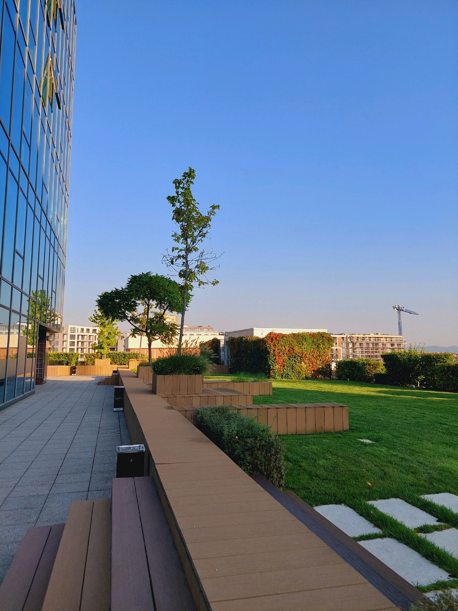 An outdoor area with a modern building on the left, featuring a glass facade. The area includes a tiled walkway leading to a wooden-decked seating area. Beyond this, there is a well-maintained green lawn with square planters and trees. In the background, there are residential buildings under construction, and a construction crane is visible against a clear blue sky. The scene appears to be a rooftop garden or a terrace with urban landscaping. The location is identified as Sofia, Bulgaria. There are no people visible in the image.