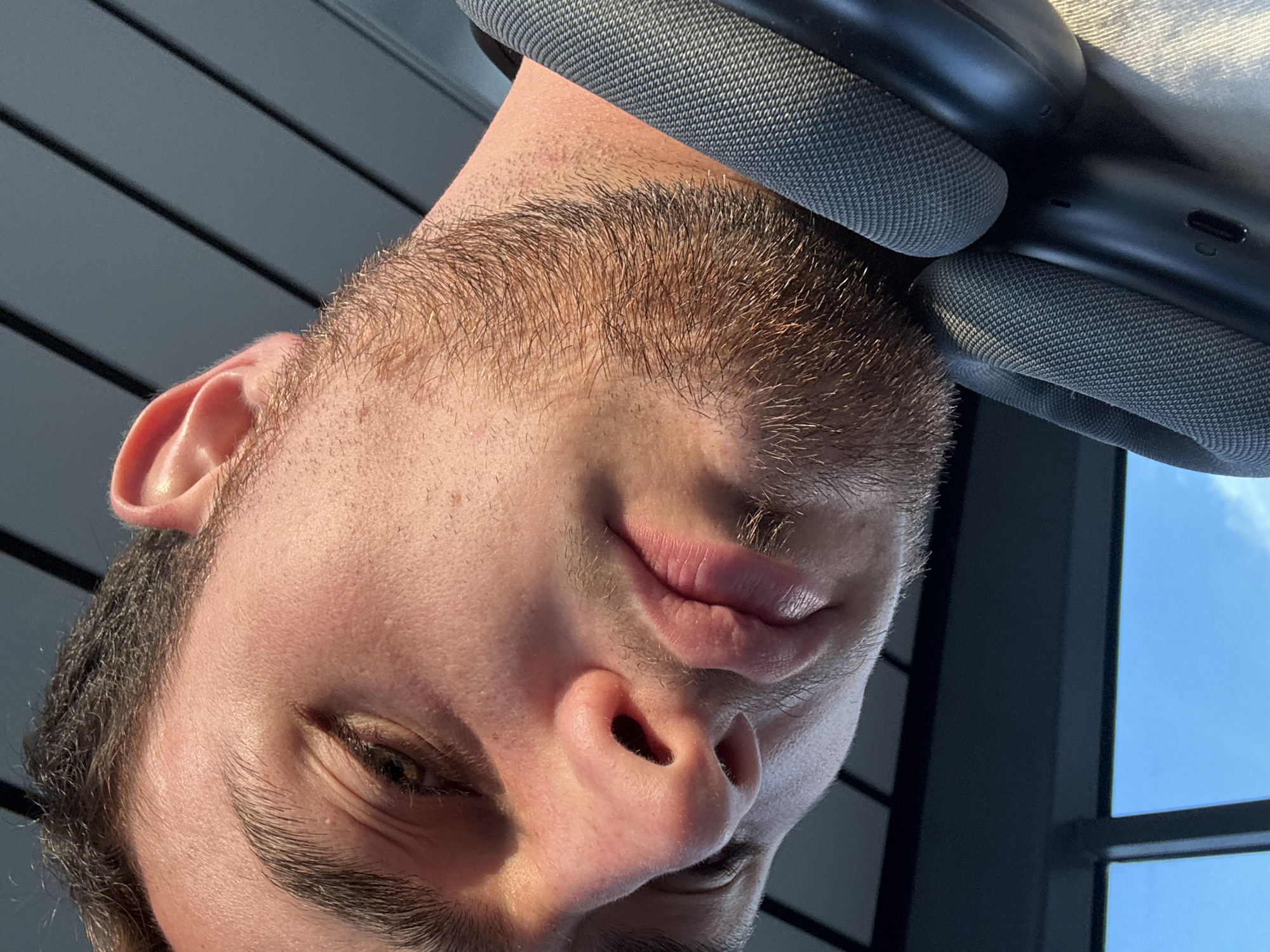 A close-up, low-angle shot features a young man's face, angled downwards, with his eyes partially closed. He has light brown hair, light skin, and a light stubble beard. The man is wearing dark gray headphones around his neck. The background consists of dark horizontal lines, suggestive of blinds or slats, and a bright blue sky with scattered clouds visible through a window. The lighting suggests it is daytime, with sunlight casting shadows on the man's face. There are no identifiable landmarks or features that would indicate a specific location within Apodaca, Mexico.