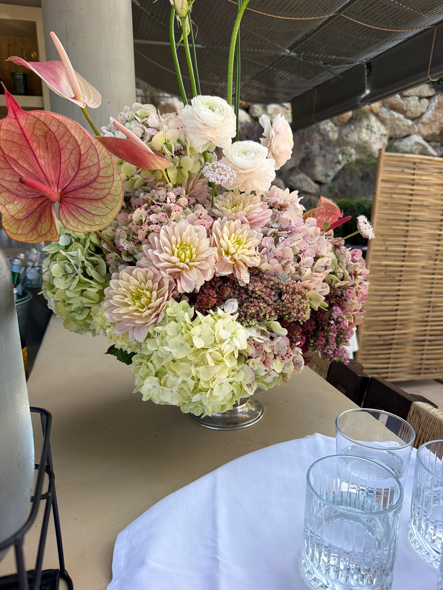 A close-up, eye-level shot captures a vibrant floral arrangement on a light-colored surface, possibly a bar or table, with a backdrop of rustic, natural elements. The arrangement, featuring a variety of flowers in shades of pink, cream, and light green, is a central focus. Prominently displayed are large pink anthurium flowers with intricate veining, interspersed with soft pink ranunculus, dahlias, and clusters of hydrangeas and other small blossoms. The bouquet is housed in a silver-toned vase. To the right of the floral arrangement, three clear, ridged glasses filled with a clear liquid are positioned on a white cloth. In the background, a woven bamboo screen and a textured stone wall suggest an outdoor or semi-outdoor setting. The overhead structure is a dark, textured material, possibly a pergola or awning, with visible ropes. The overall impression is one of an outdoor event or gathering, possibly in a location like Sant Josep de sa Talaia, Spain, known for its natural beauty and resort-style venues. No people are visible in the frame.
