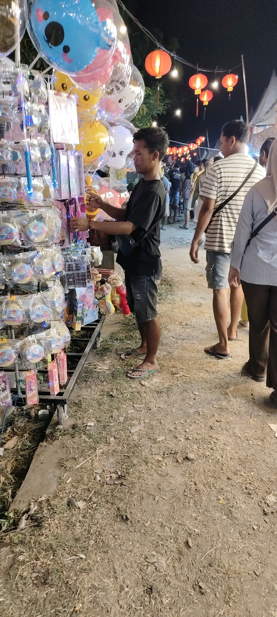 In Pulo, Indonesia, a night market scene unfolds. A young man, wearing a black t-shirt and denim shorts, stands at a stall filled with various items. He appears to be arranging or inspecting some of the merchandise, which includes brightly colored balloons, some with cartoonish faces, and other packaged goods displayed on shelves. The stall is illuminated with blue lights.
In the background, several other individuals are visible, some walking through the market. Red lanterns are strung across the scene, casting a warm glow and indicating a festive atmosphere. A white tent structure is also present in the distance. The ground is covered with dirt and dry grass, suggesting an outdoor setting. The overall impression is that of a lively local market at night.
