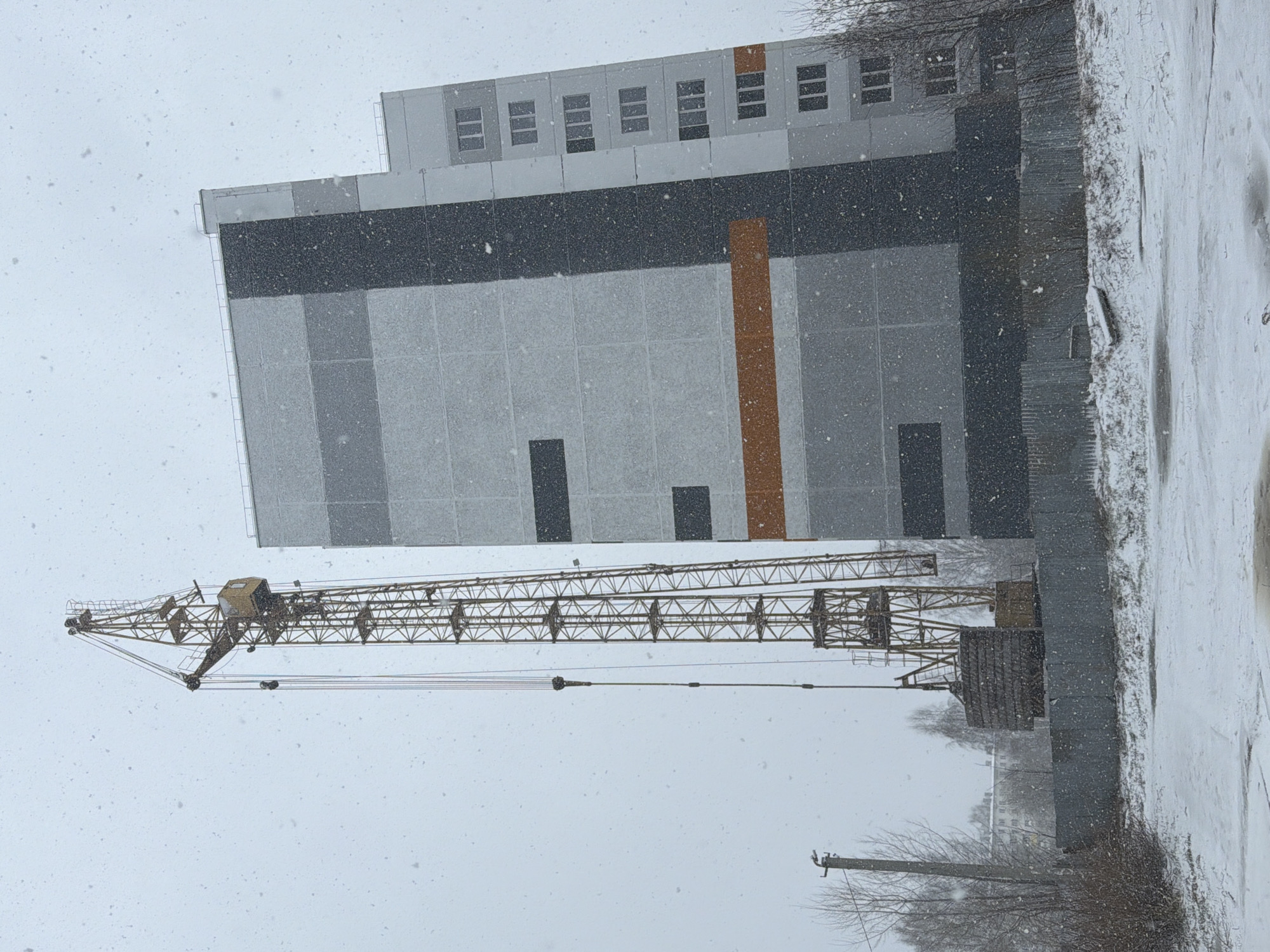 The image depicts a snowy, overcast scene in Chaikovsky, Russia. A construction crane is positioned in front of a multi-story building under construction. The building's facade is a mix of gray and dark gray panels, with a vertical orange stripe. Windows are visible on the upper floors. Snow covers the ground and is actively falling. A metal fence runs along the right side of the frame. Bare trees are visible in the background.