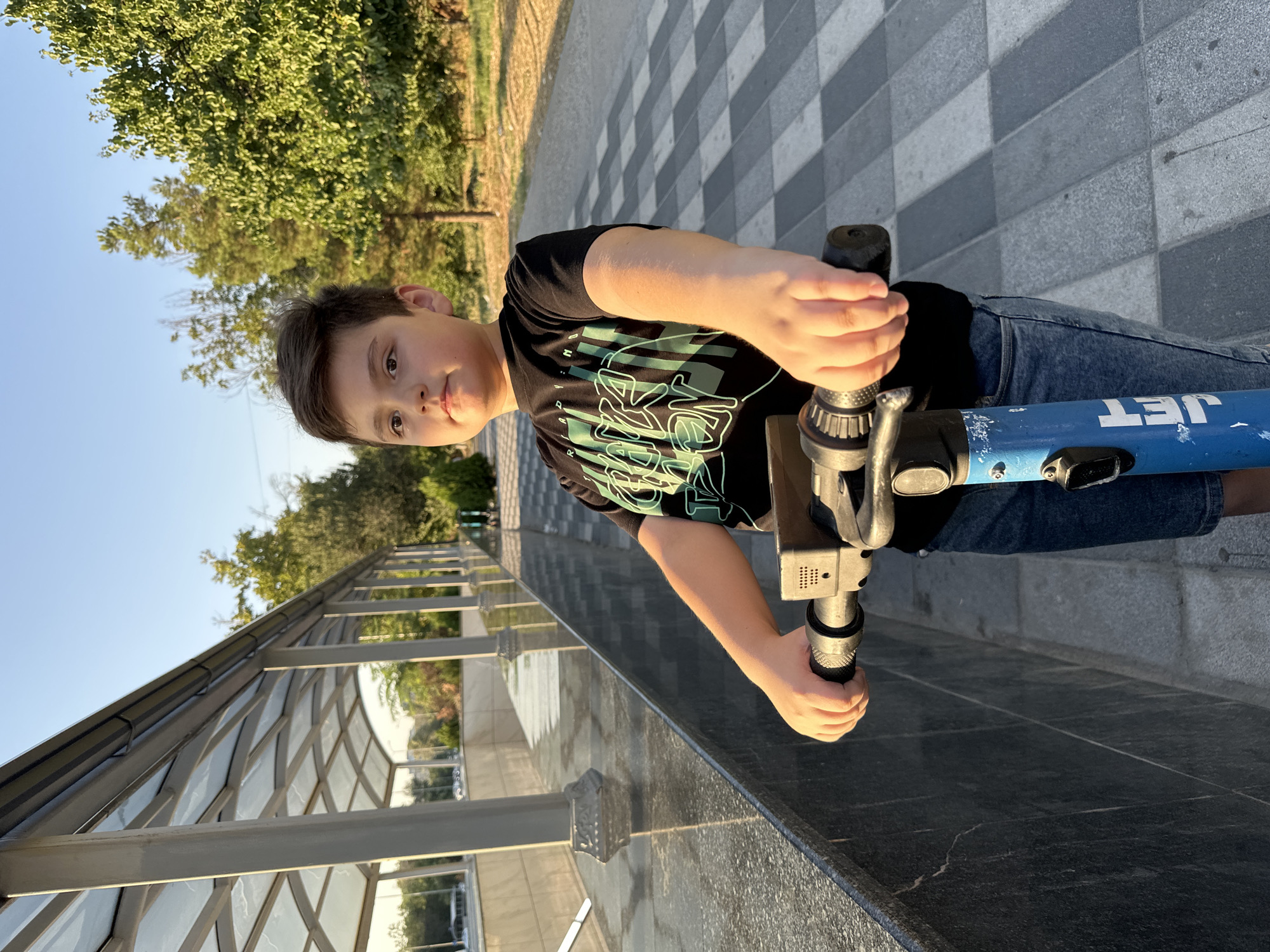 A young boy, appearing to be around 8-10 years old, stands on a blue electric scooter on a paved walkway. He is wearing a black t-shirt with green text and denim shorts. The boy is looking slightly to his right with a neutral expression. He is holding the handlebars of the scooter with both hands. The walkway has a patterned design of grey and white squares. To the left of the walkway, there is a polished dark stone wall and a covered walkway structure with a glass roof supported by white pillars. Trees and greenery are visible in the background, and the sky is clear and blue, suggesting it is daytime. The lighting indicates it is likely late afternoon or early evening, with the sun casting a warm glow on the boy's arm. The scene appears to be a public area within the city of Tashkent, Uzbekistan, possibly near an entrance to a park or an architectural complex.